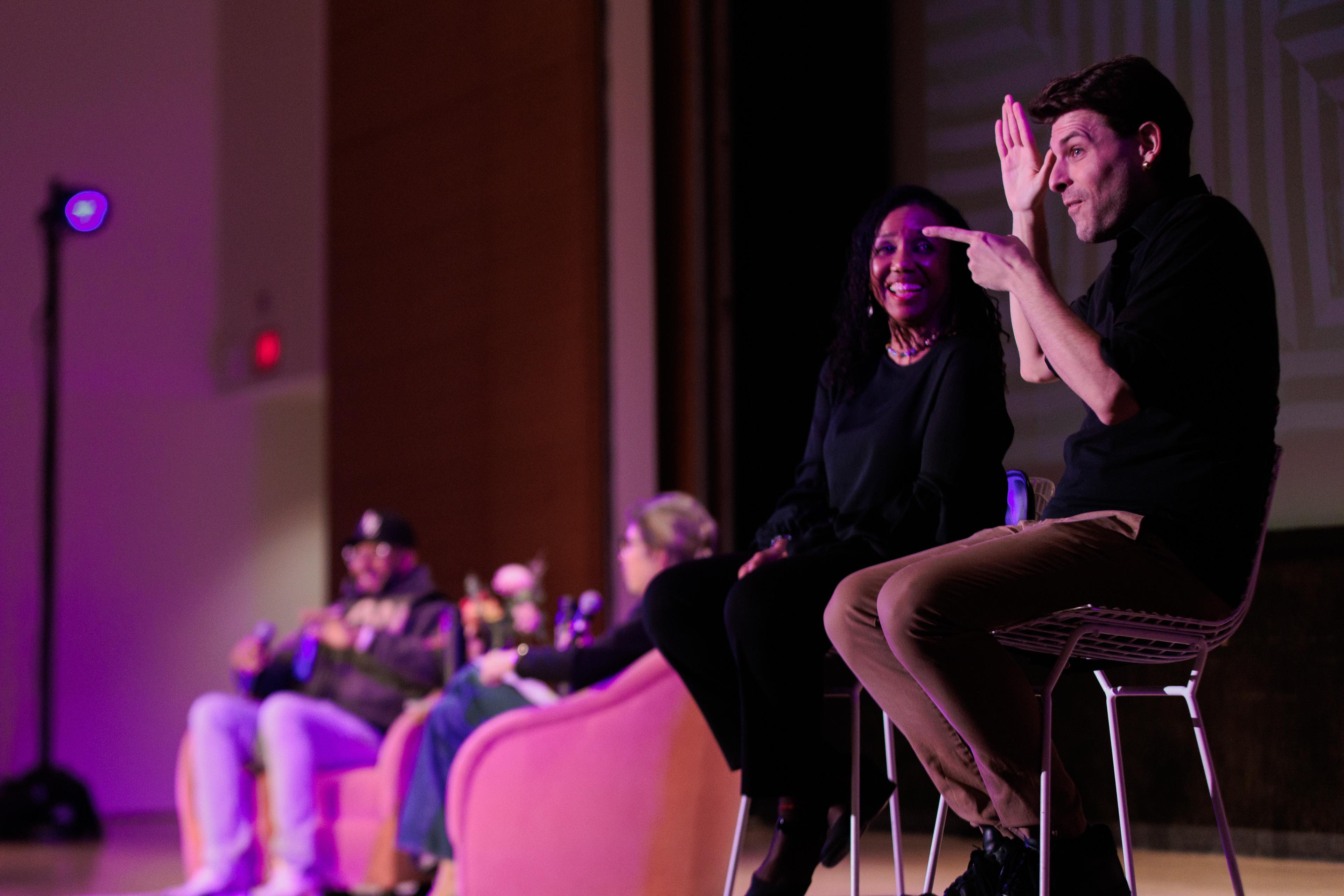 Two ASL interpretors sit on a stage, signing to the audience