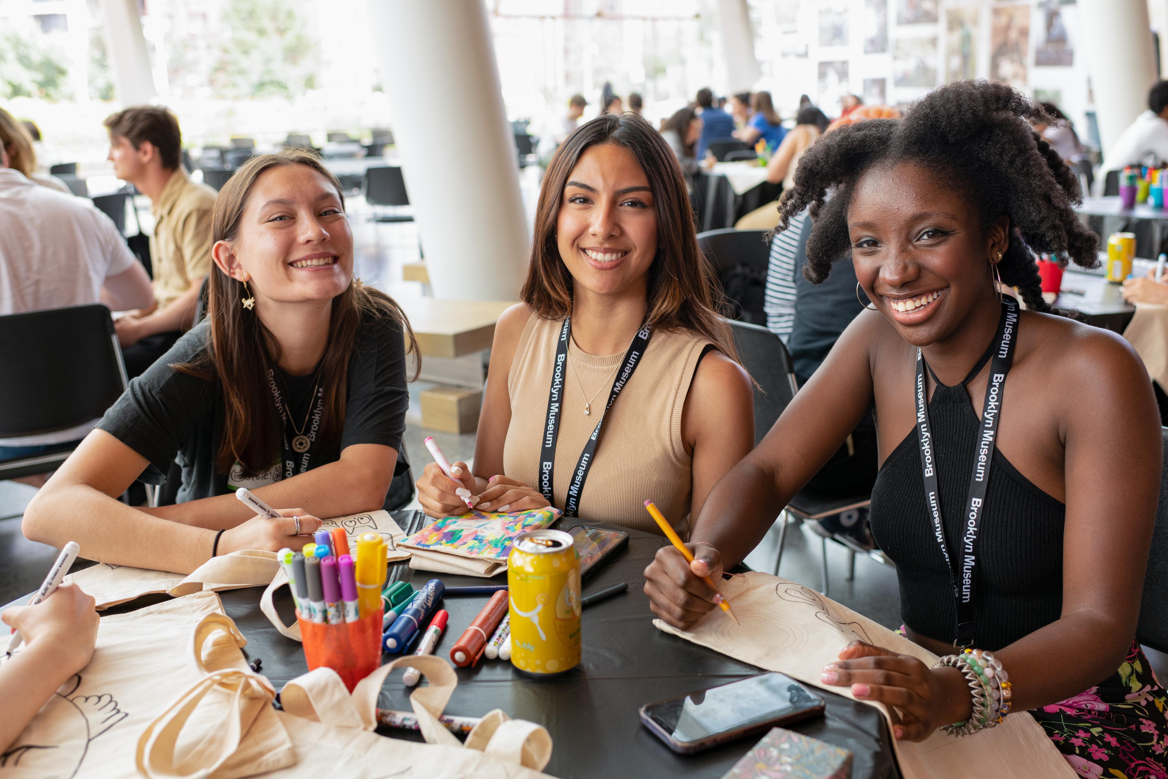 Three young adults smiling at the camera while drawing on canvas tote bags