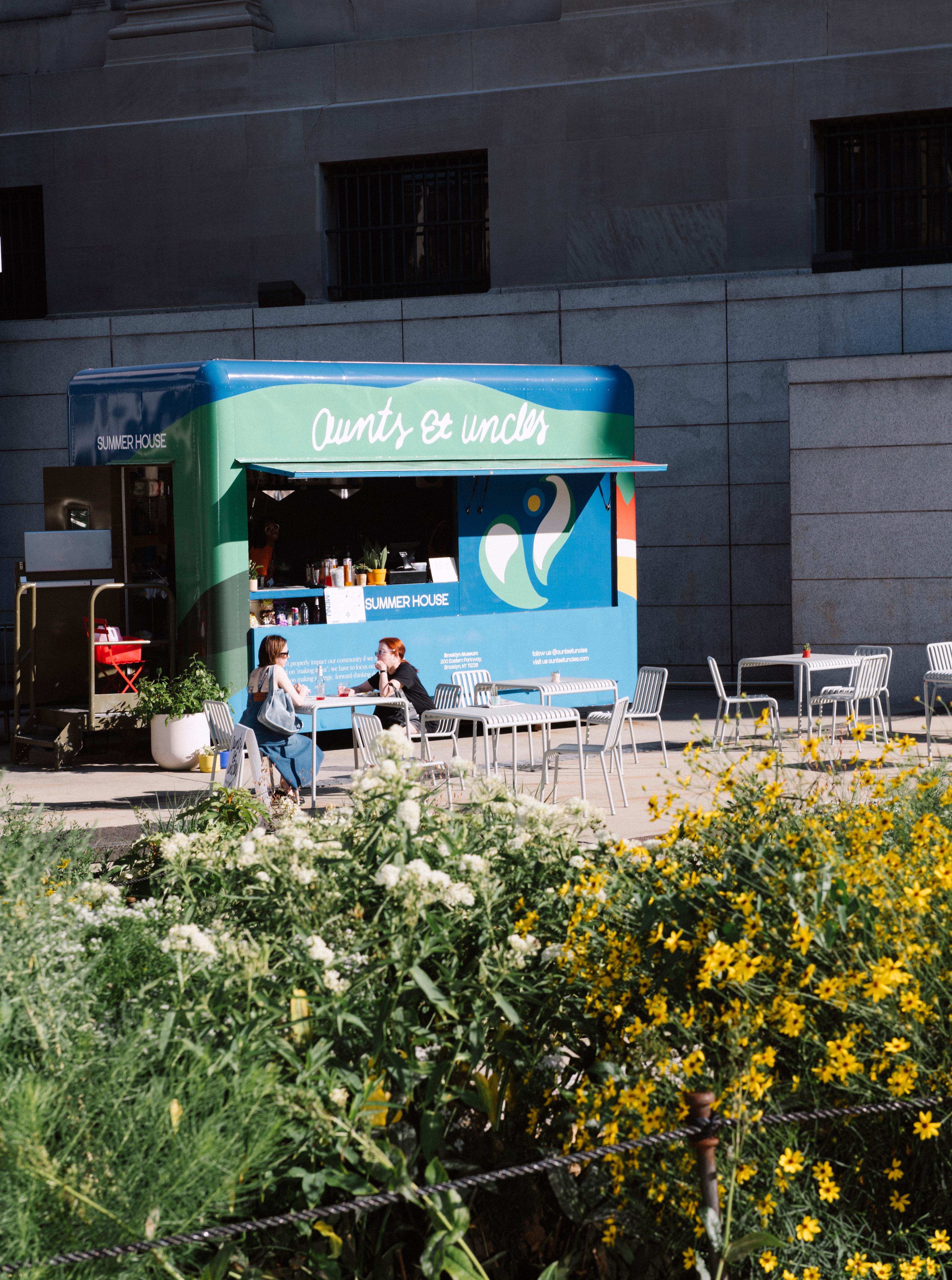 A blue and green food truck is parked against a building with two diners sitting at chairs, flowers in the foreground