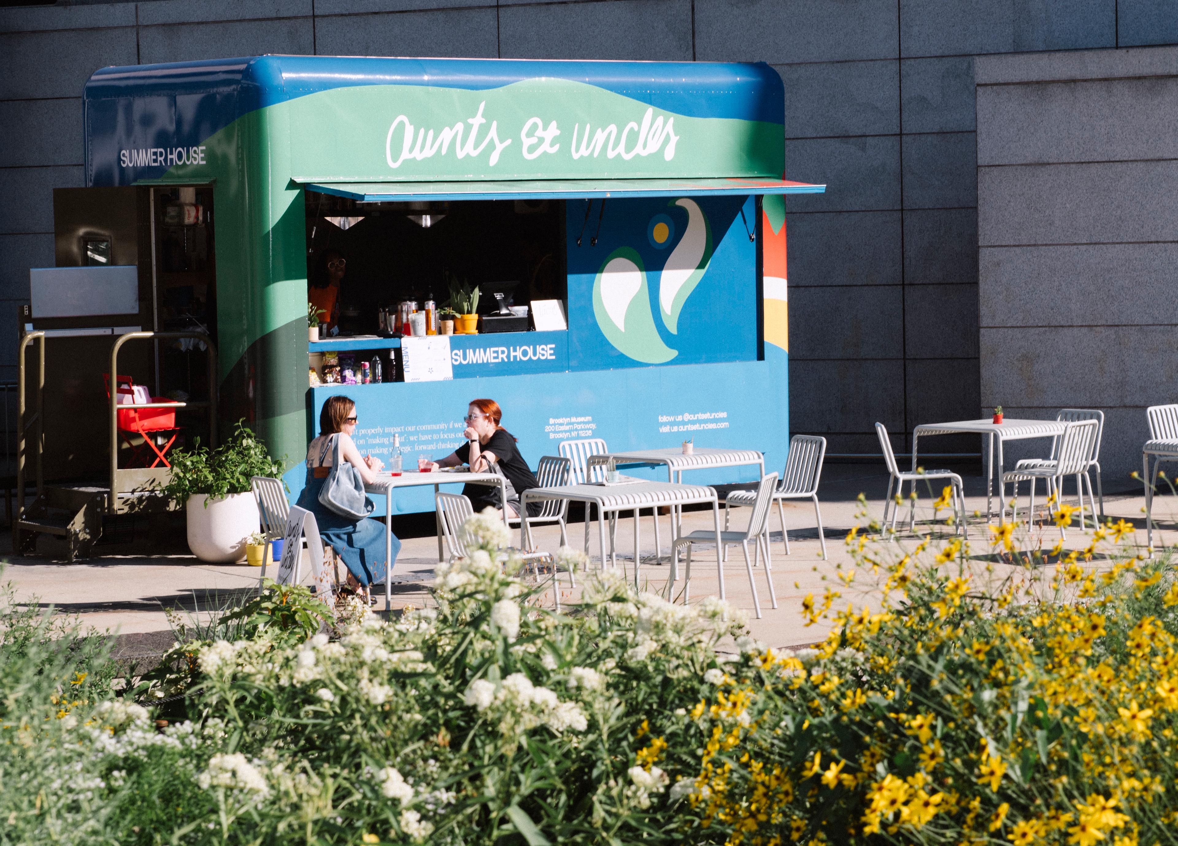A blue and green food truck is parked against a building with two diners sitting at chairs, flowers in the foreground