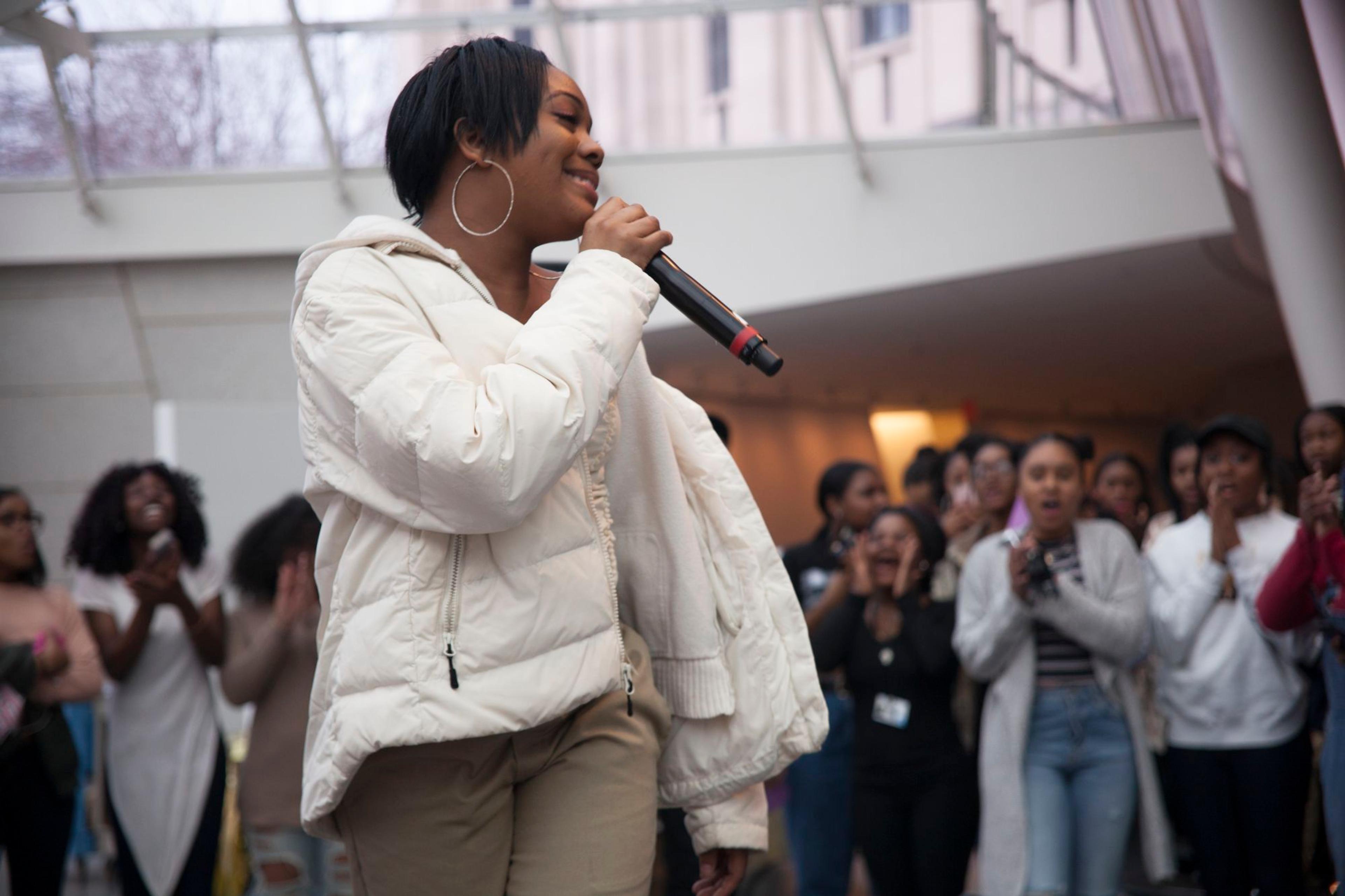 Teen in a white puffy coat smiles and sings into a mike with a crowd of peers surrounding