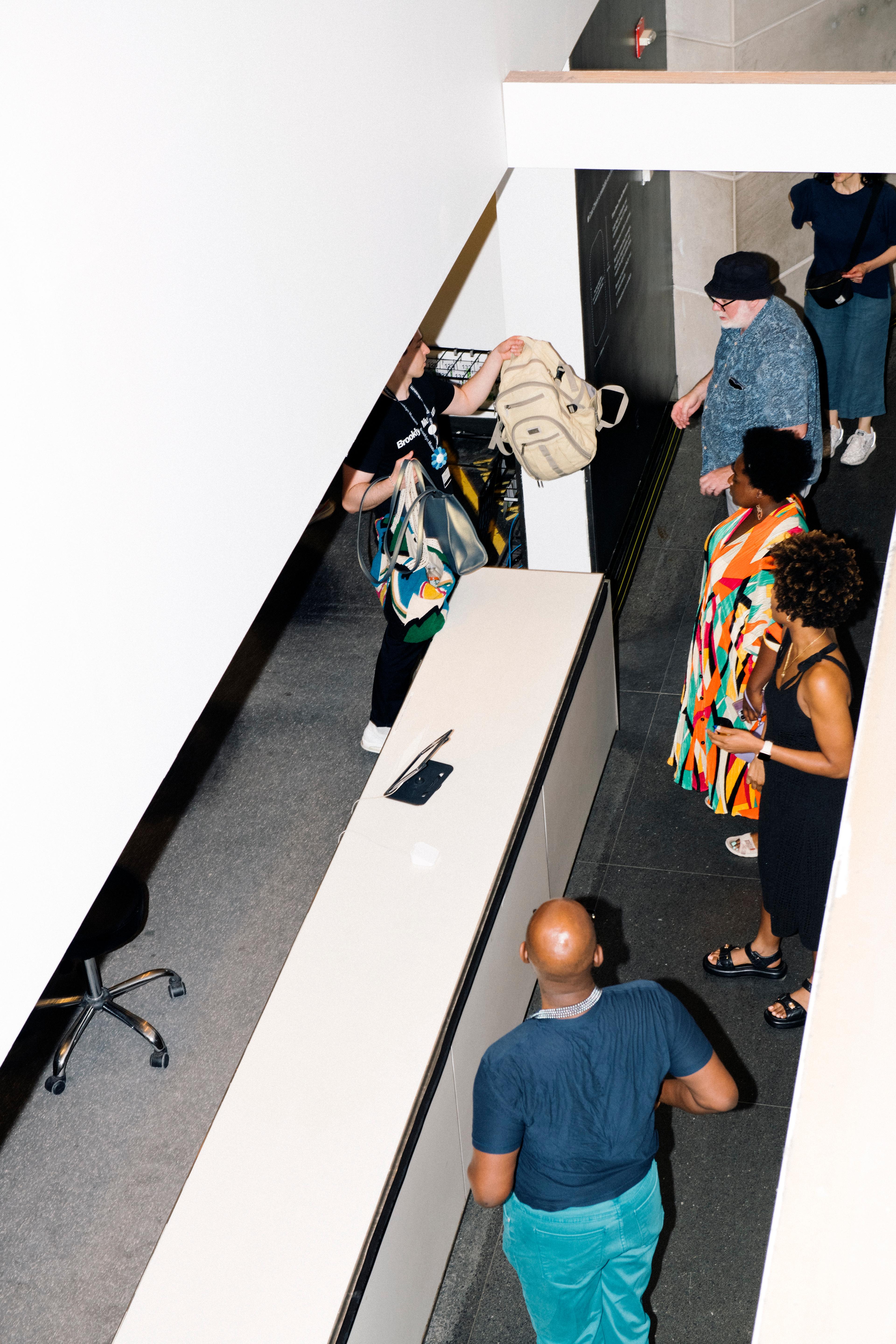 Adults line up waiting at a desk, while another adult in a black Brooklyn Museum shirt hands  one of them a bag