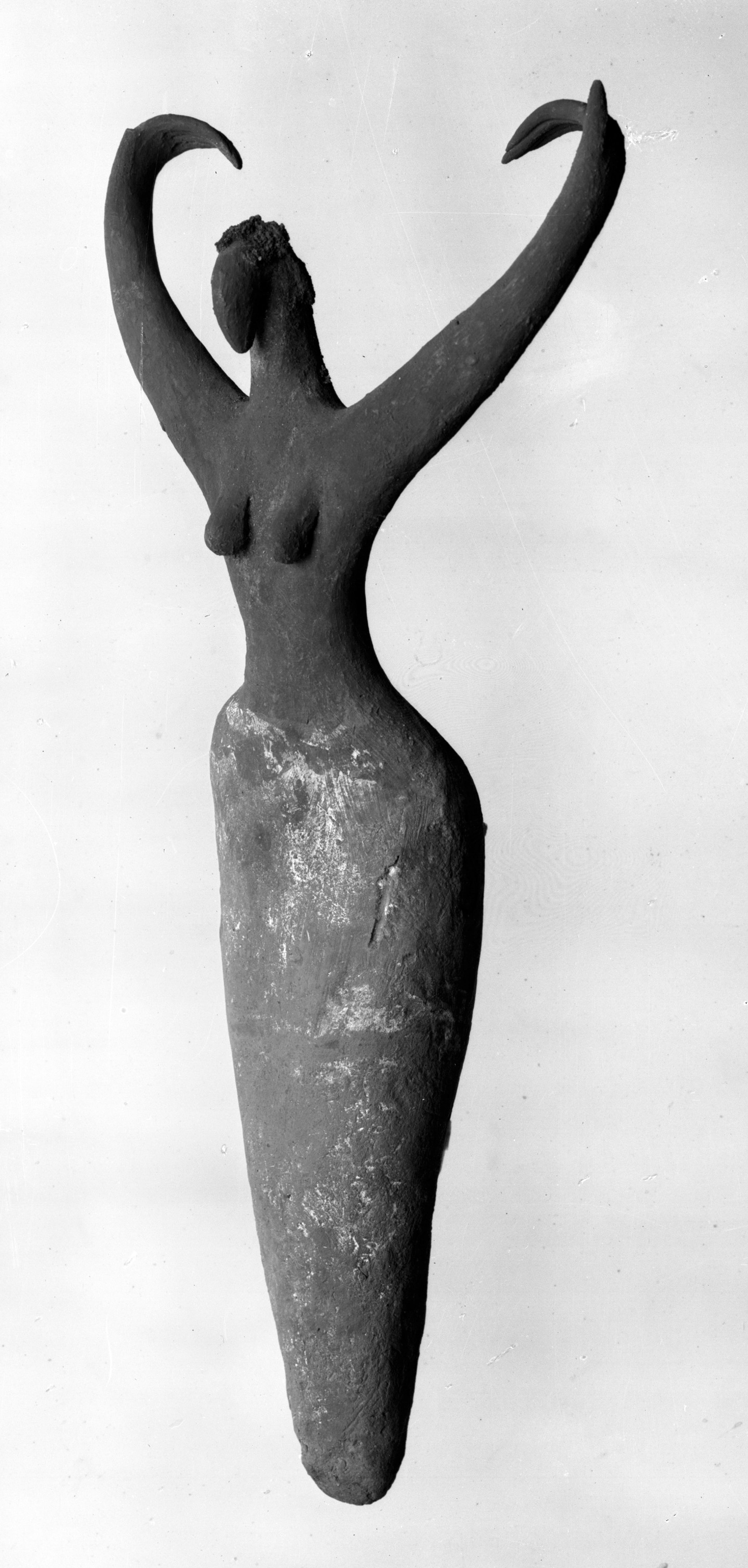 Black-and-white photograph of a sculpture of a female figure with raised arms and a tapered lower body