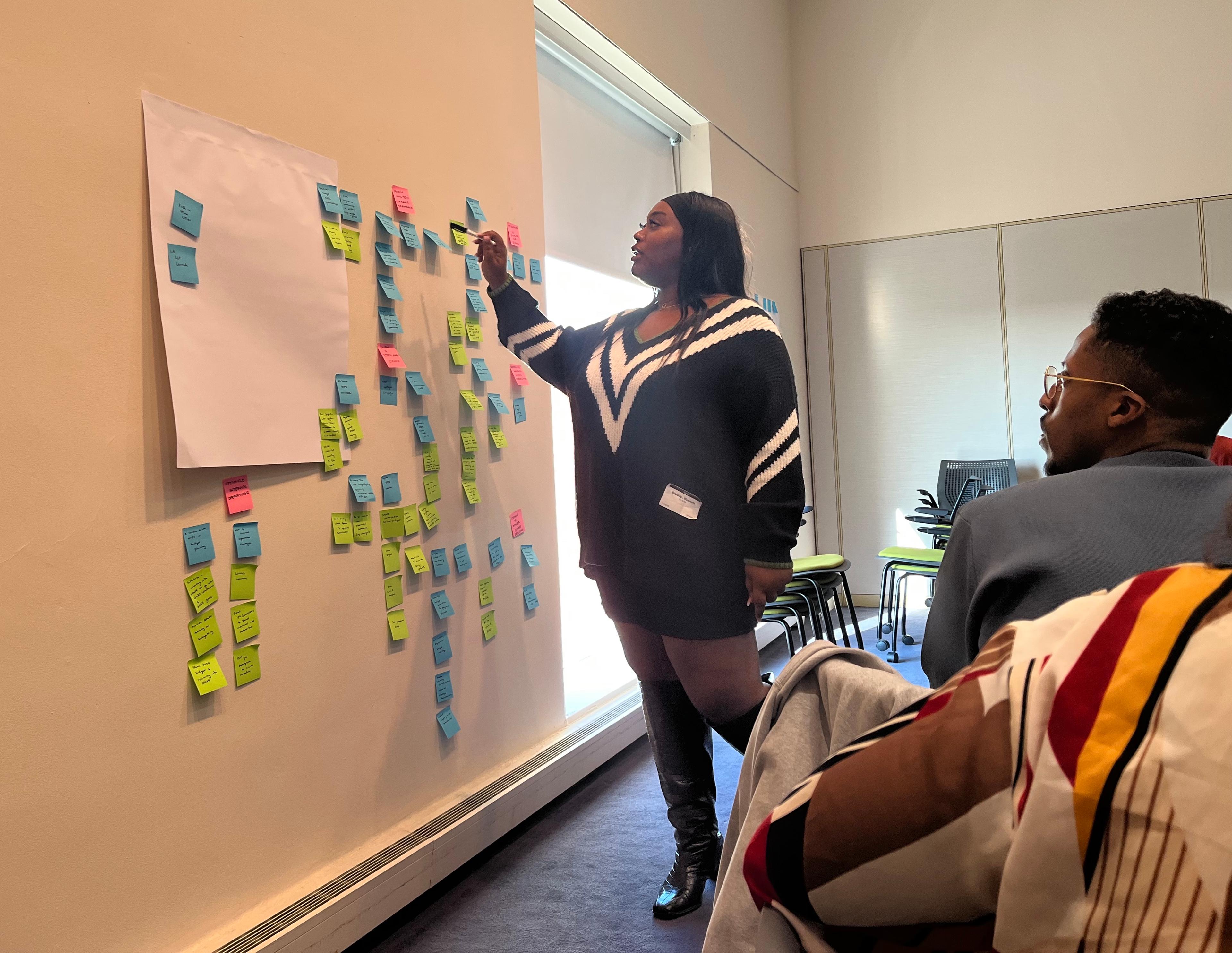 A standing by a wall of blue, green, and pink post-its and poster board, reaches for a post-it while another seated adult watches on