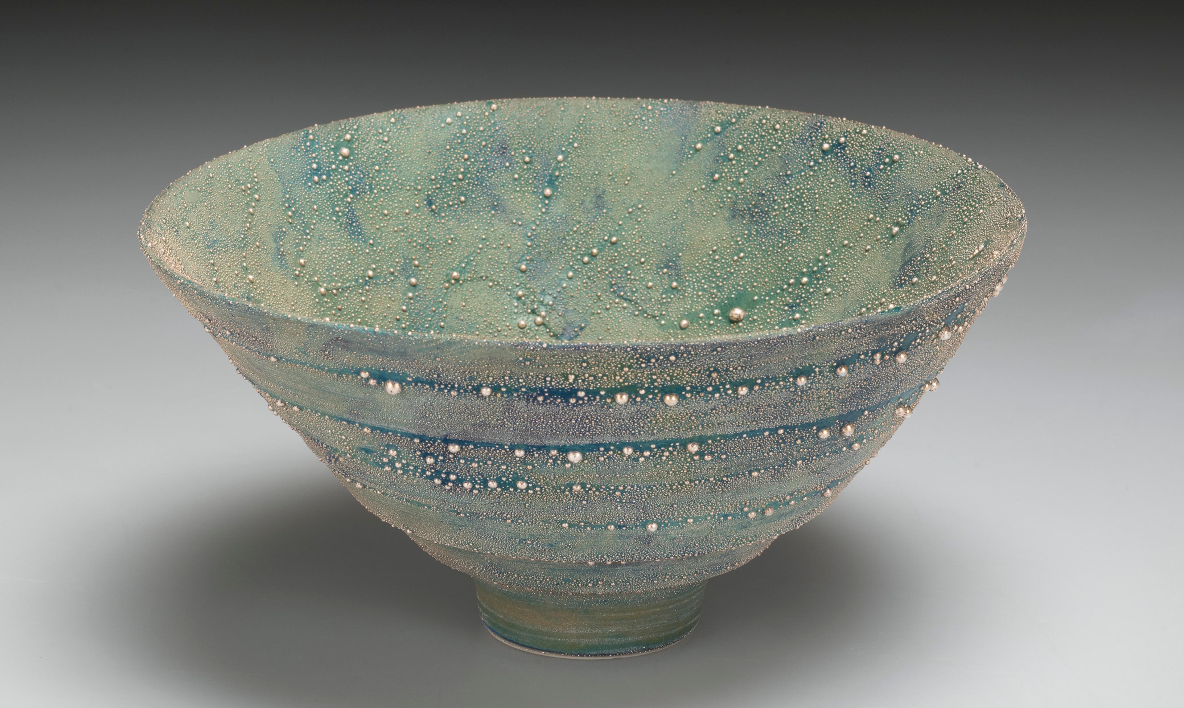 A ceramic bowl, the green surface is mottled with splashes of blue and covered with beads of silver