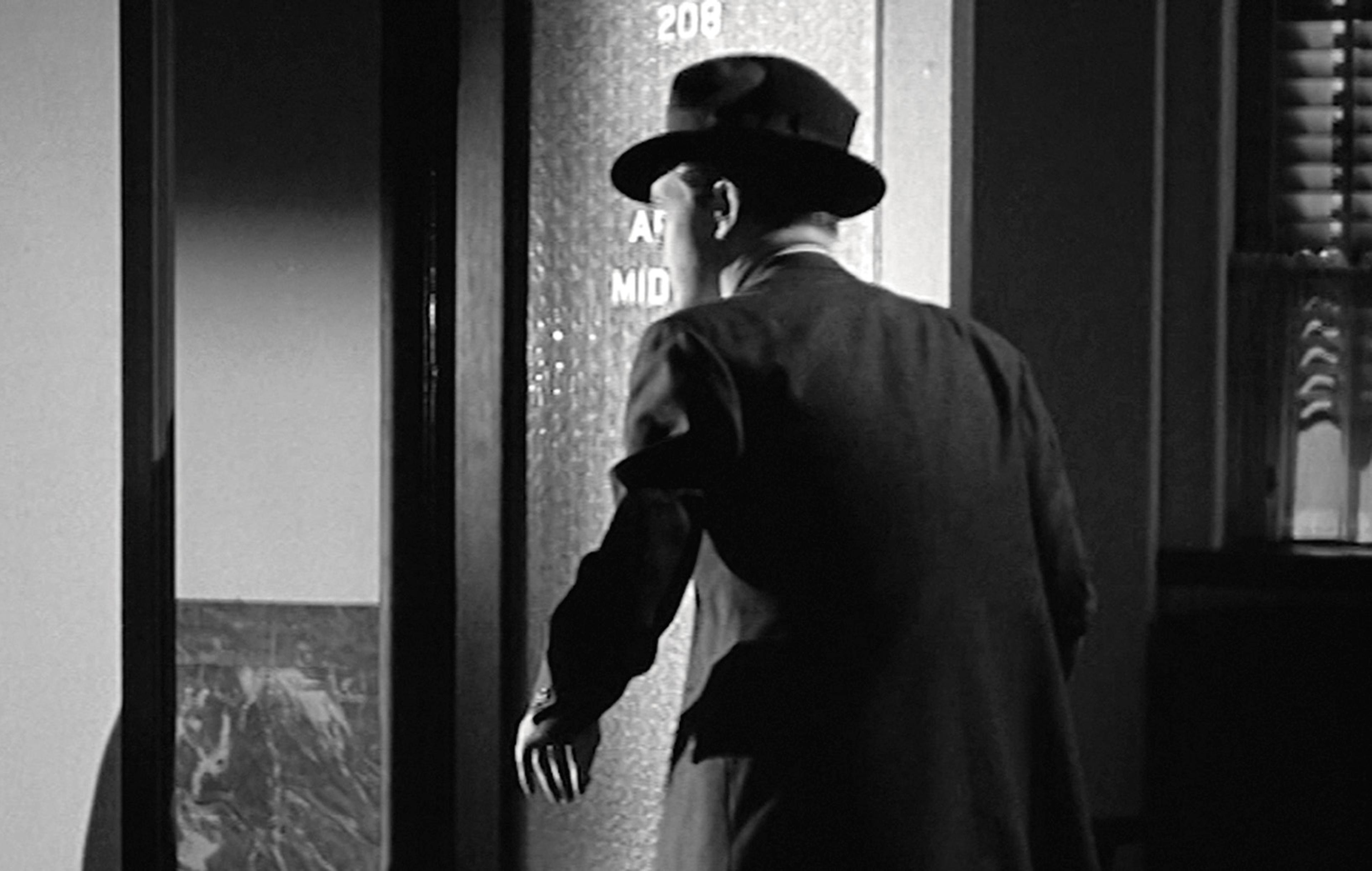 Film still showing a man in a hat looking through a doorway