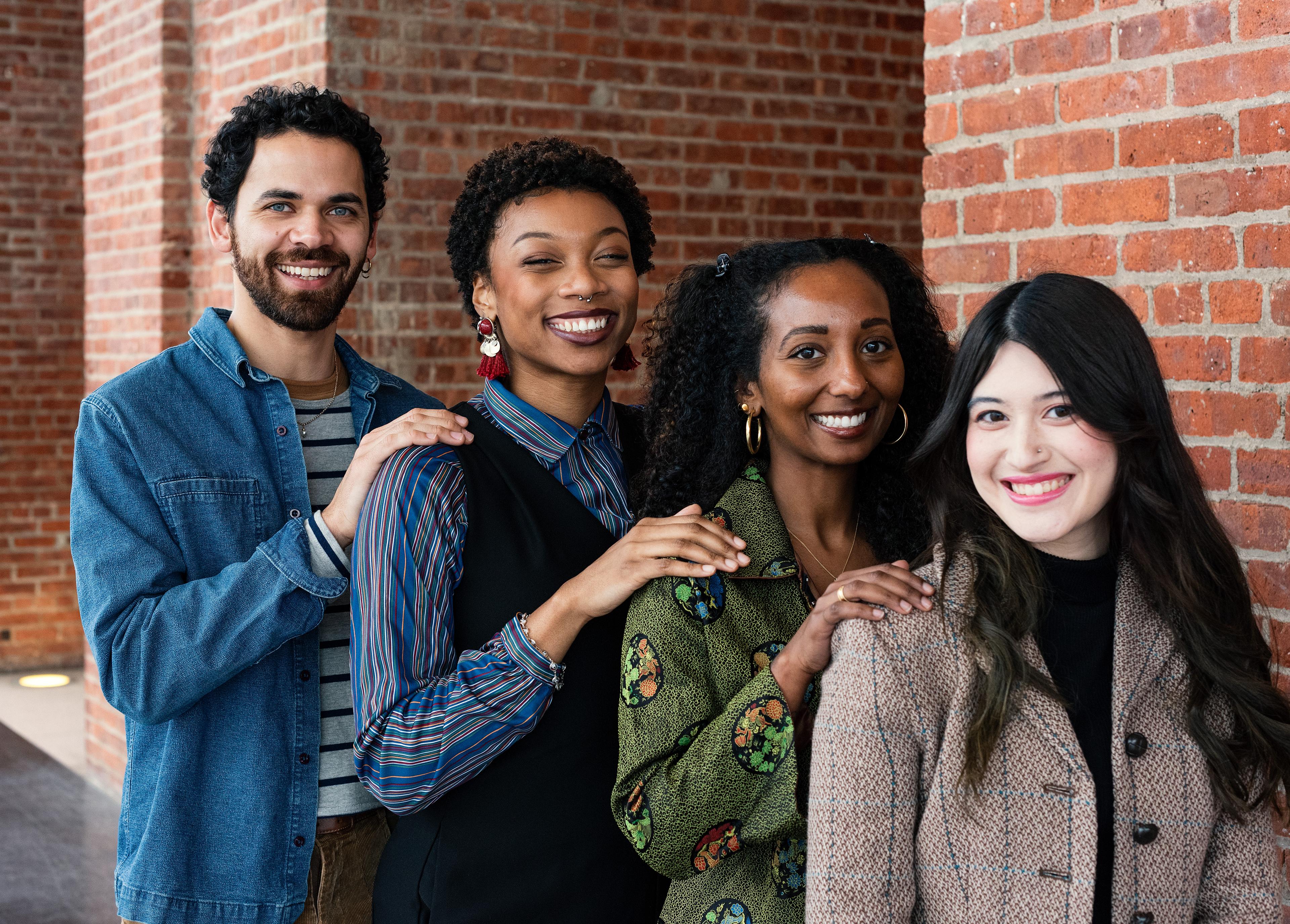 Four young adults stand lined up in order of height, smiling with their hands on eachother's shoulders