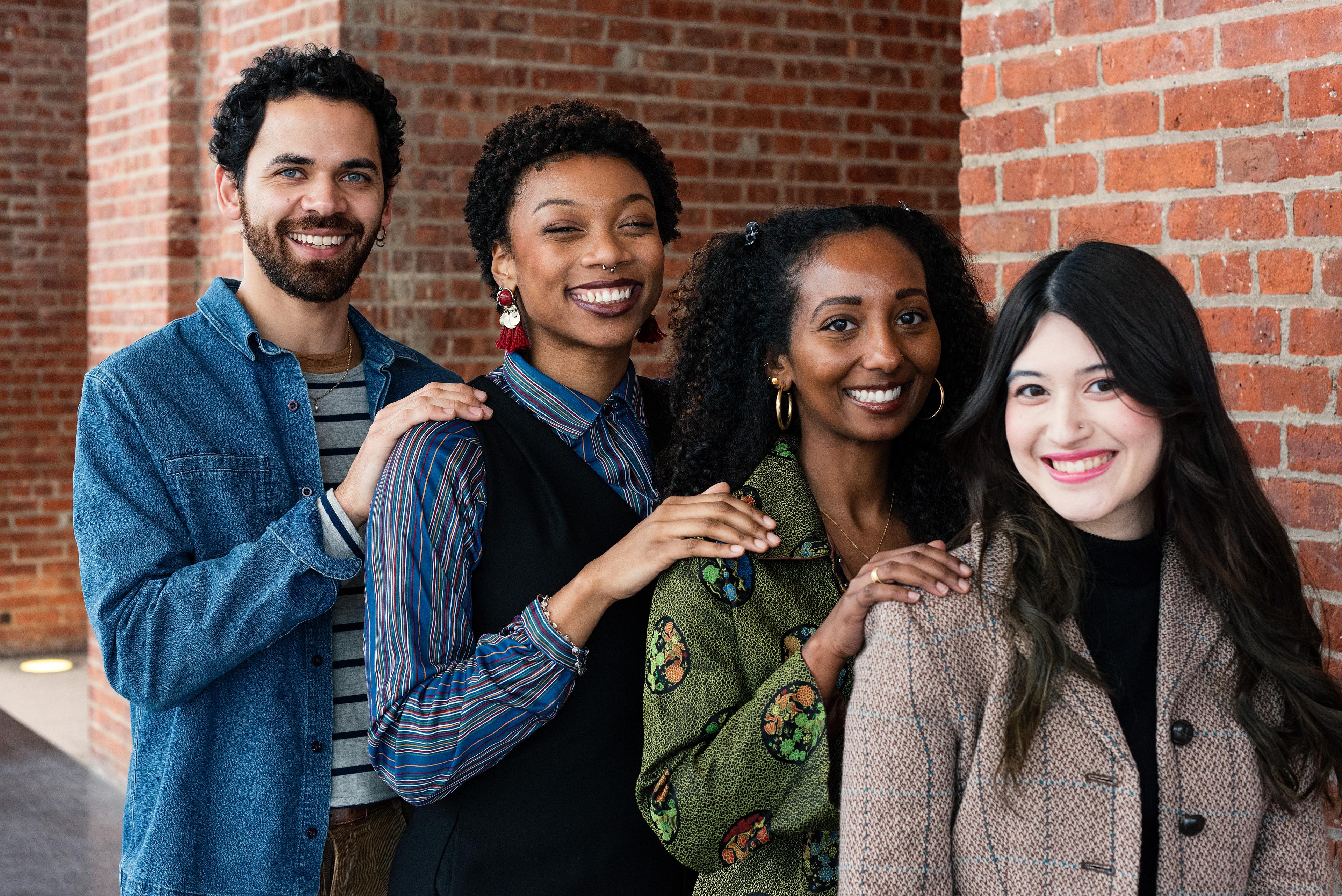 Four young adults stand lined up in order of height, smiling with their hands on eachother's shoulders