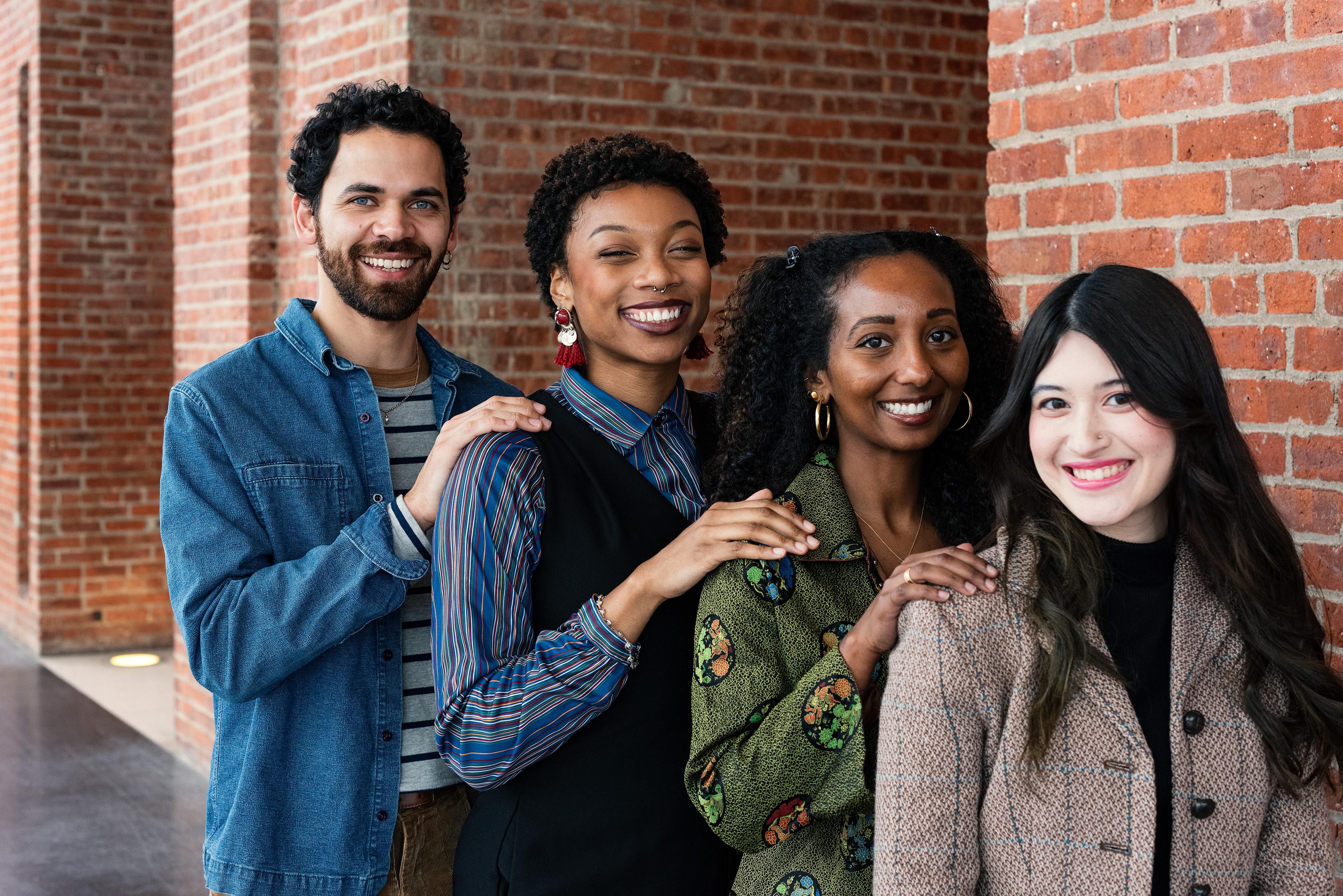 Four young adults stand lined up in order of height, smiling with their hands on eachother's shoulders