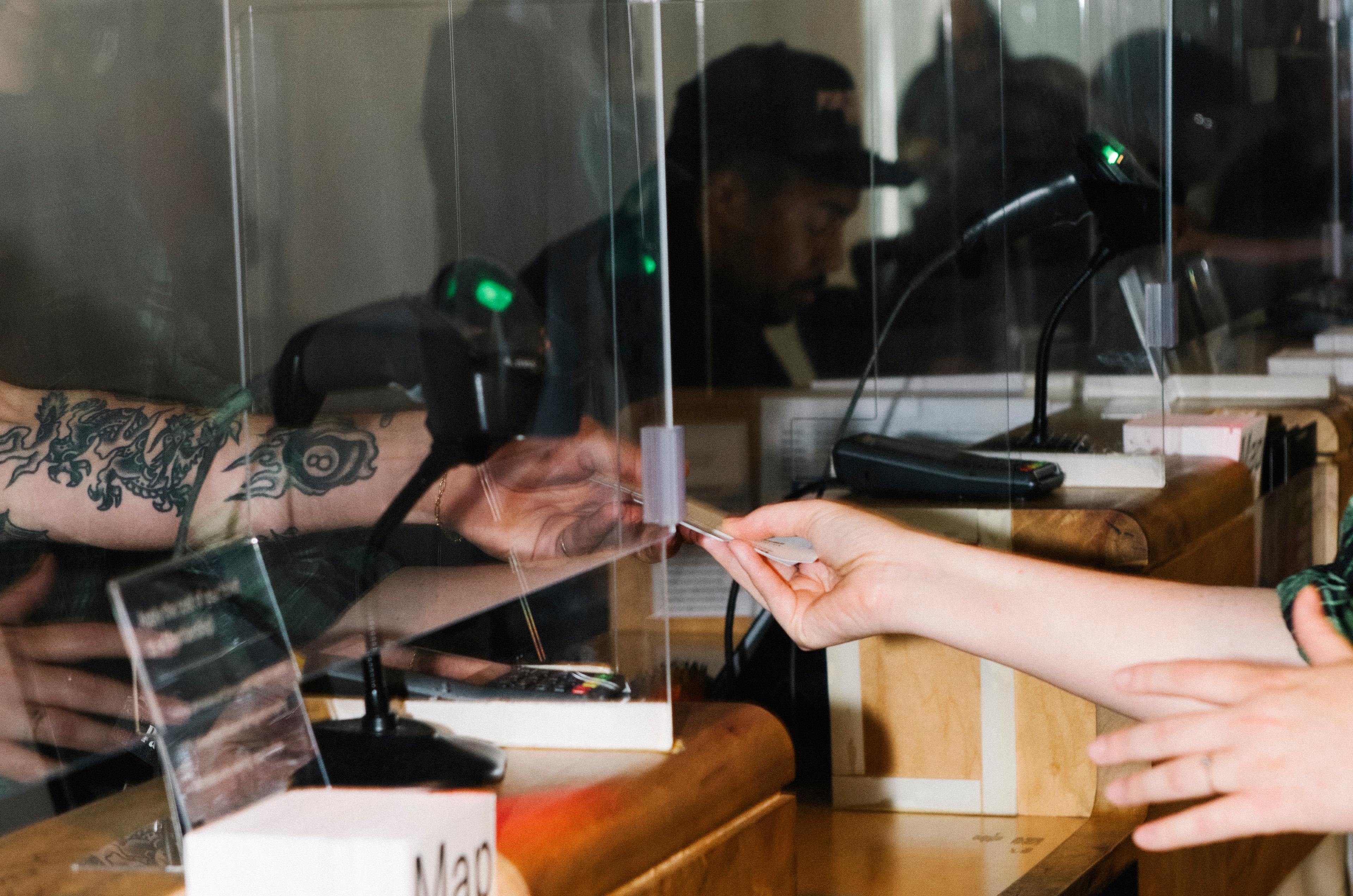 Detail of tattooed arm handing a ticket to another hand through a glass window that reads "Member Services"