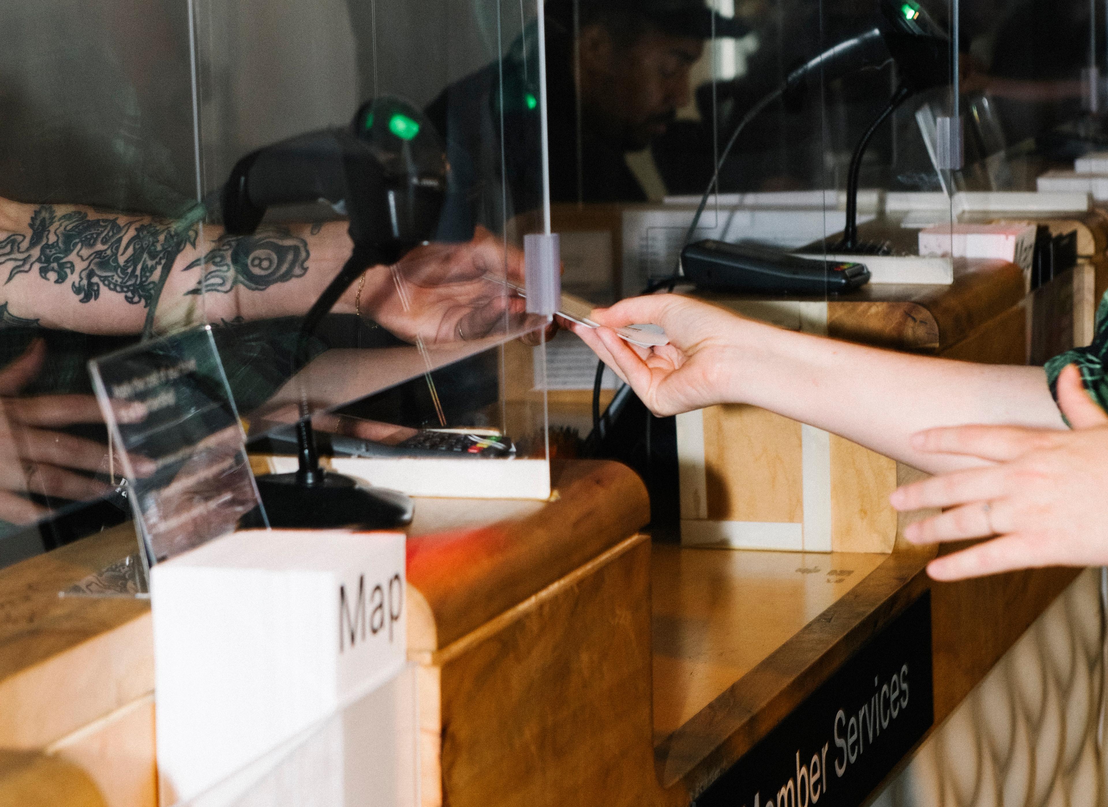 Detail of tattooed arm handing a ticket to another hand through a glass window that reads "Member Services"