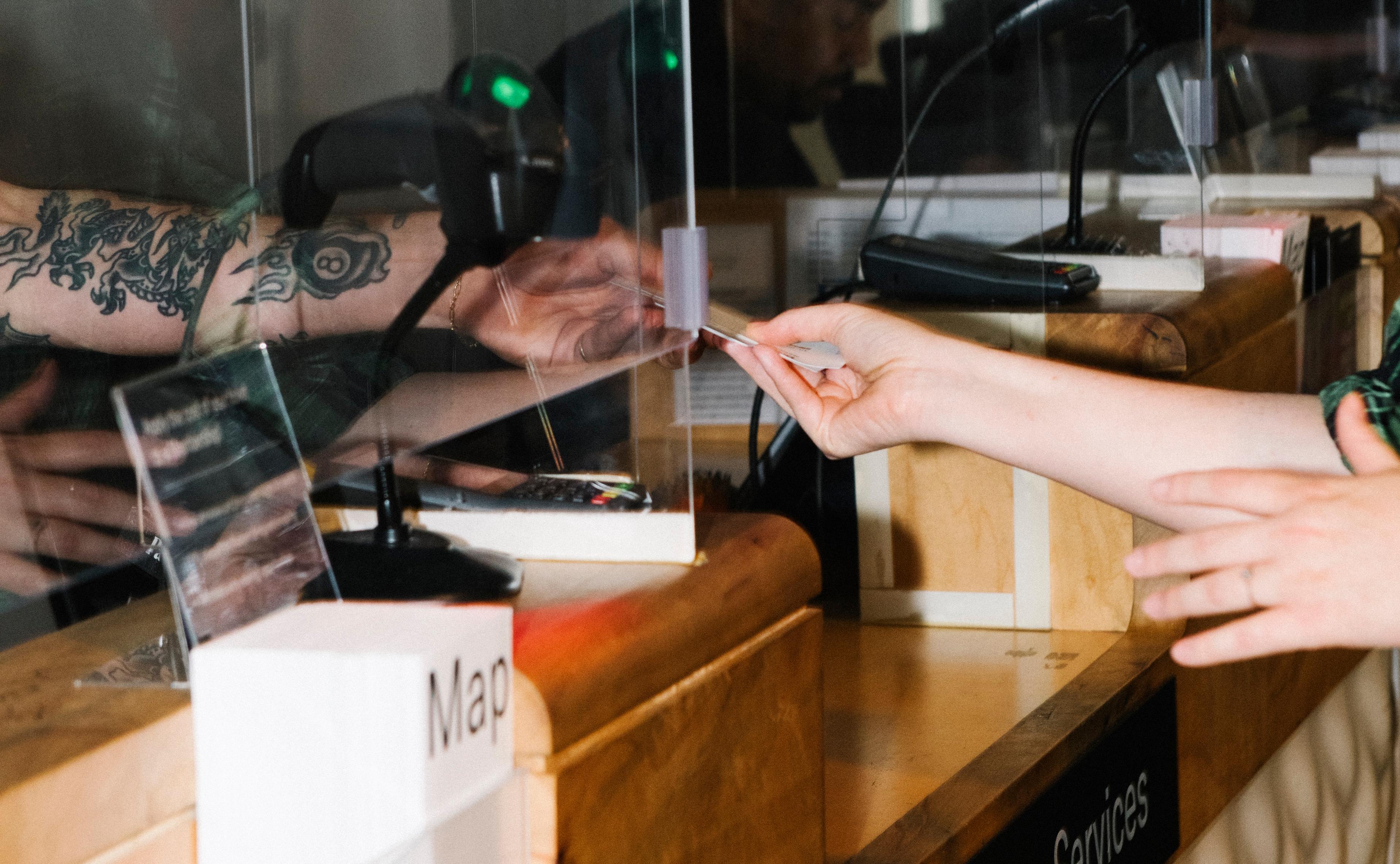 Detail of tattooed arm handing a ticket to another hand through a glass window that reads "Member Services"