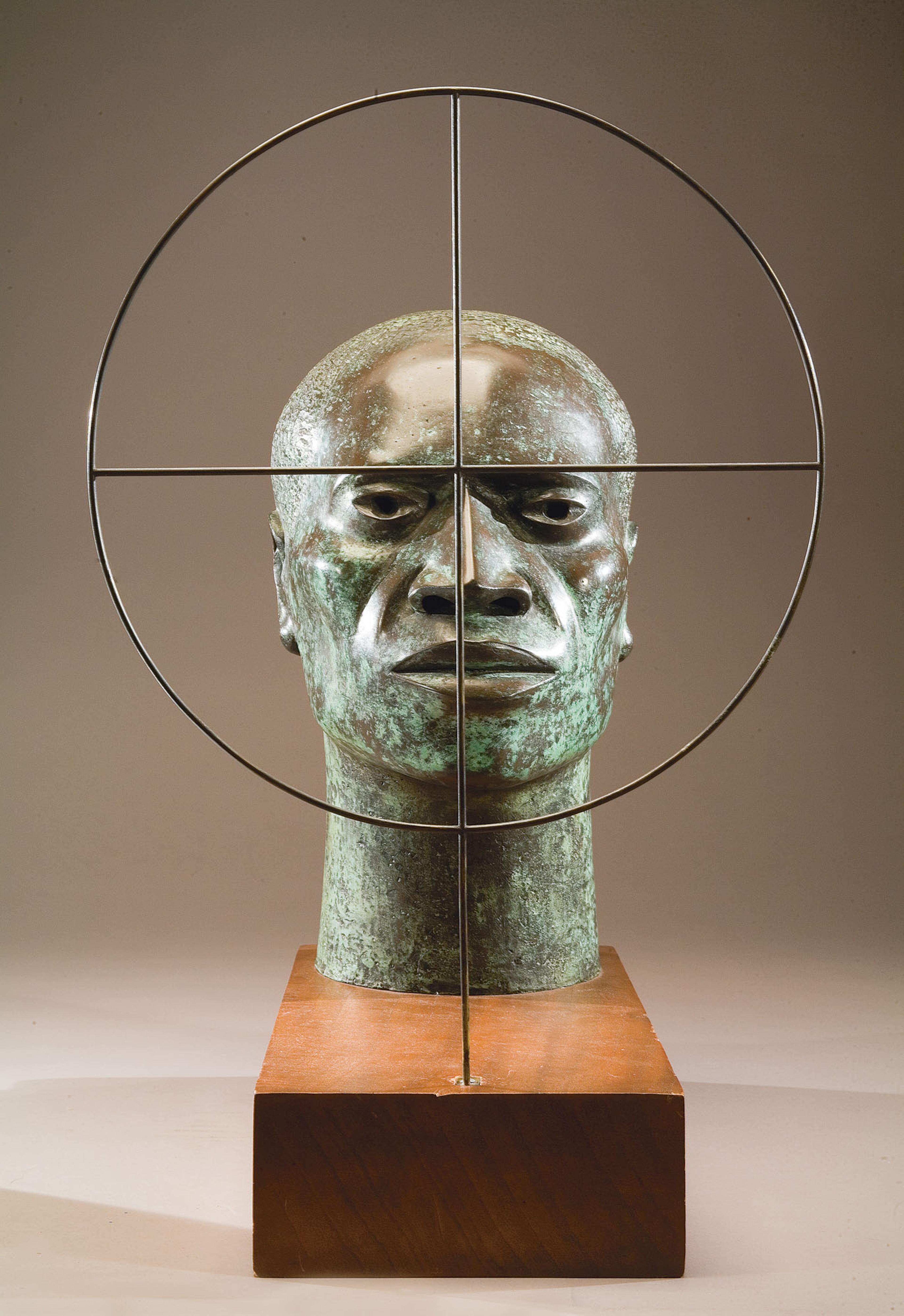 Sculpture of a bronze head on a wooden base, set behind a metal target, against a grey background