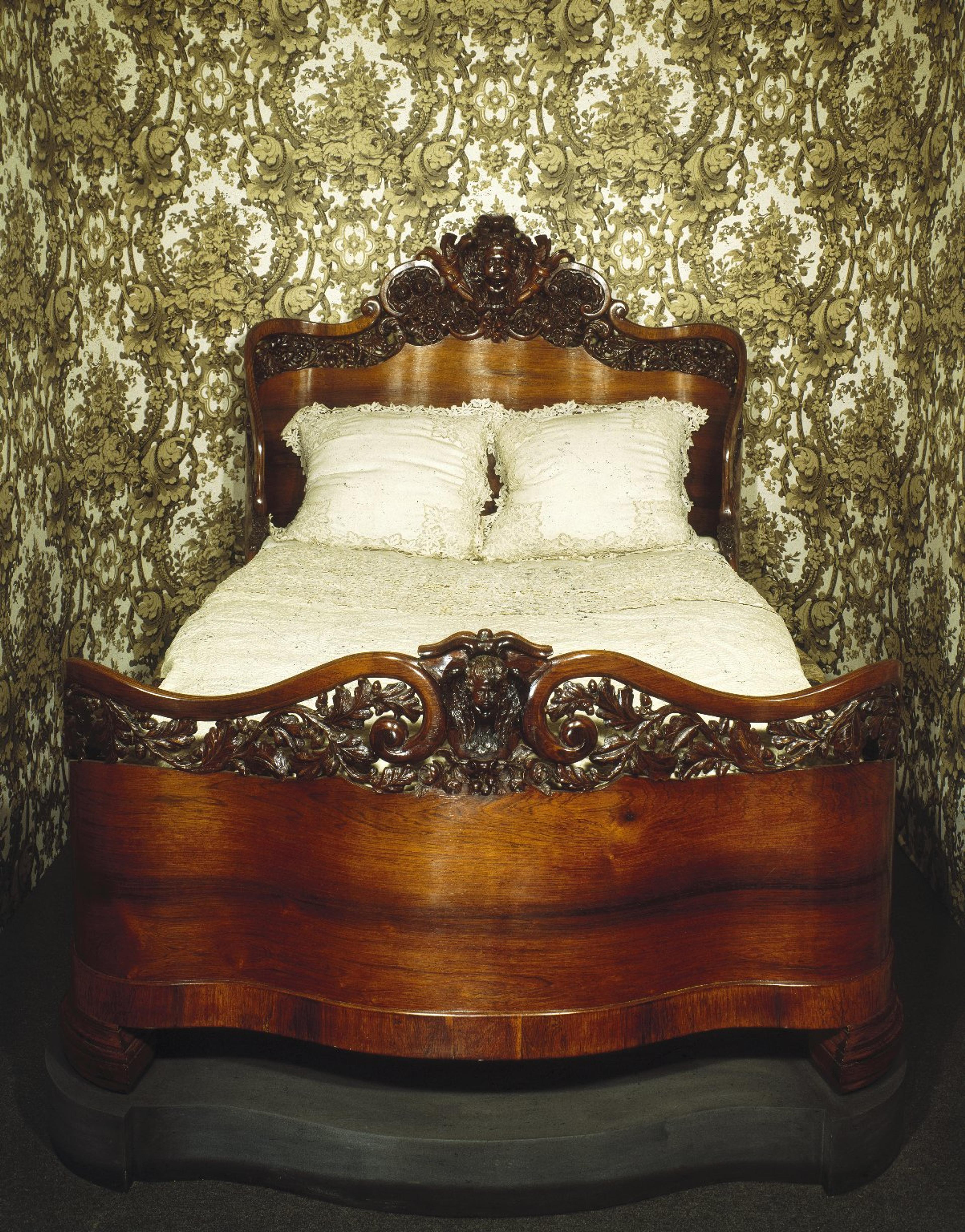 A Victorian-style bed with elaborate carved wood details and ornate floral wallpaper