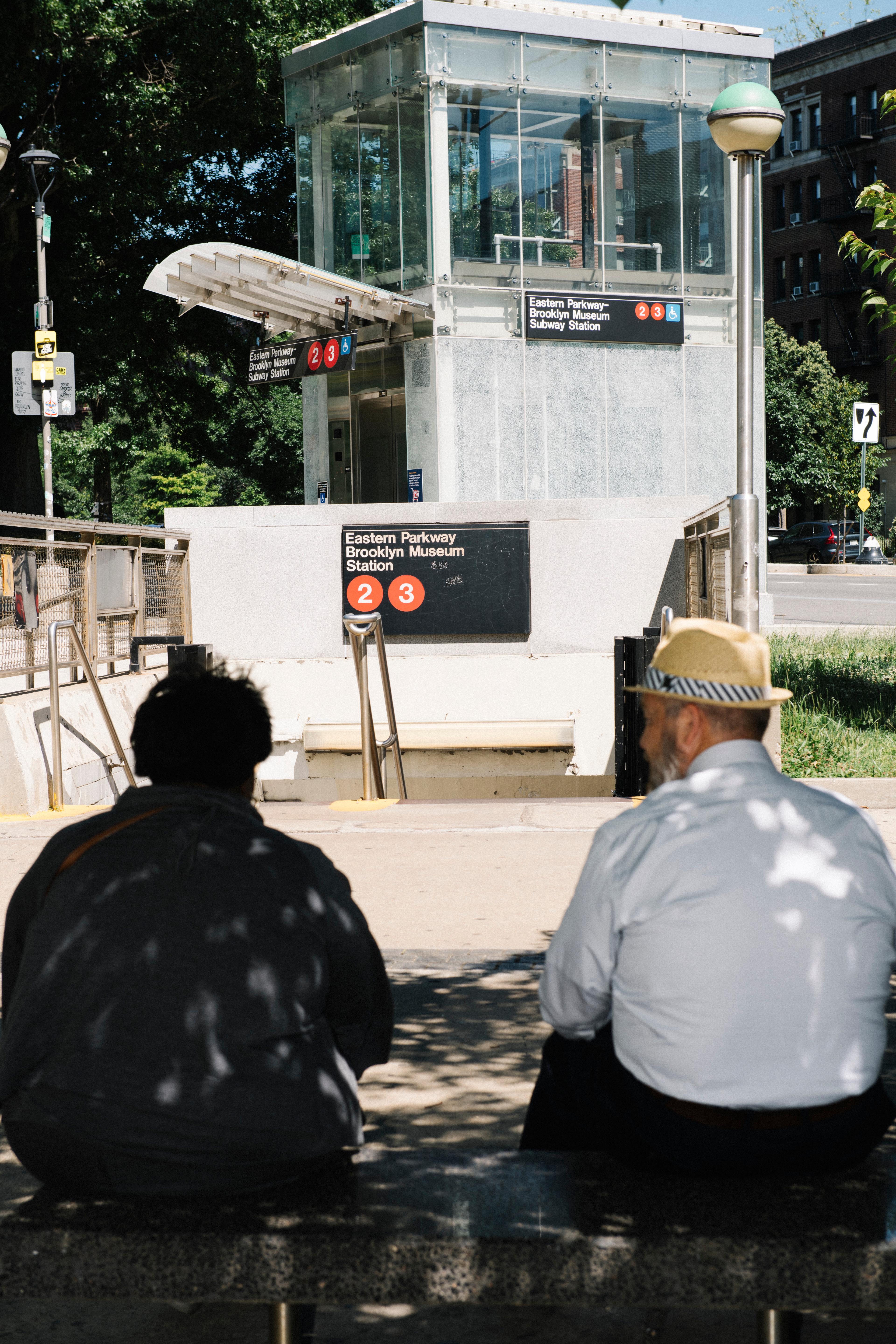 Two adults seated in on a bench in the shade face a subway elevator entrance that reads "Eastern Parkway Brooklyn Museum Station 2 3"