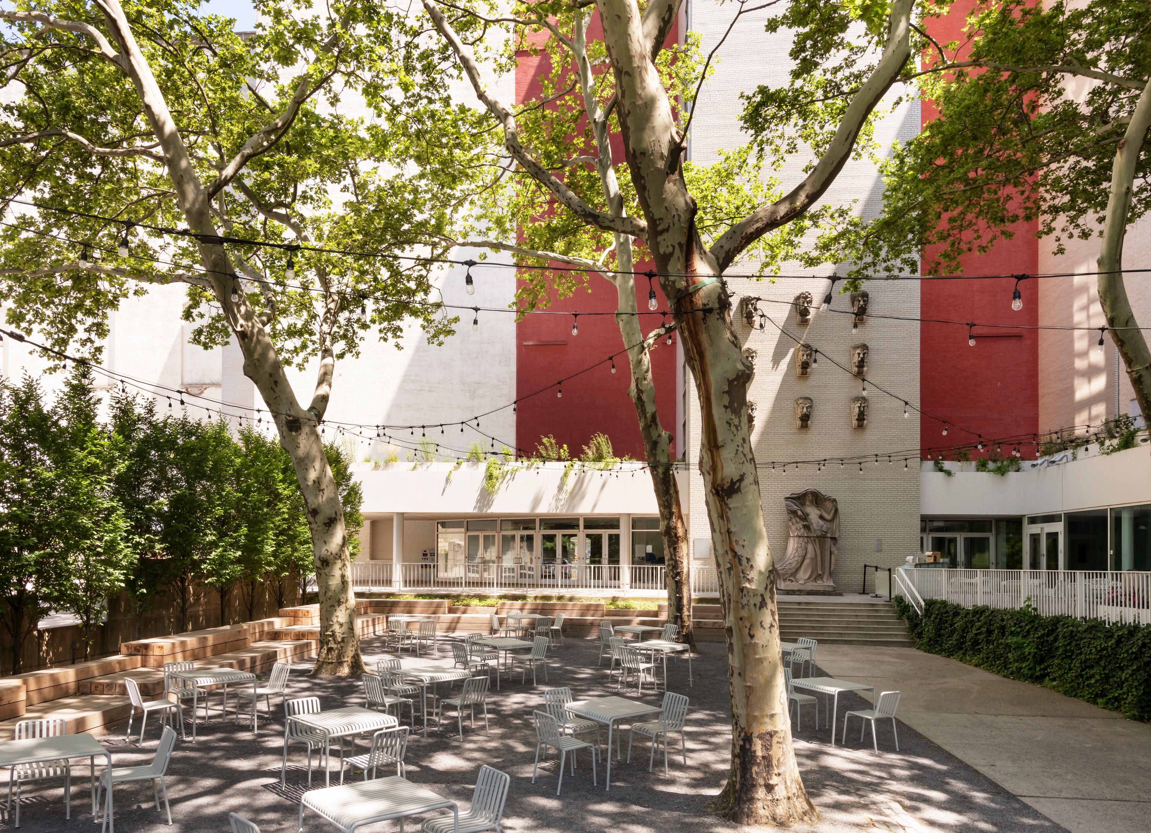 Tables and chairs arranged in the shade of trees with the back entrance to the Brooklyn Museum in background