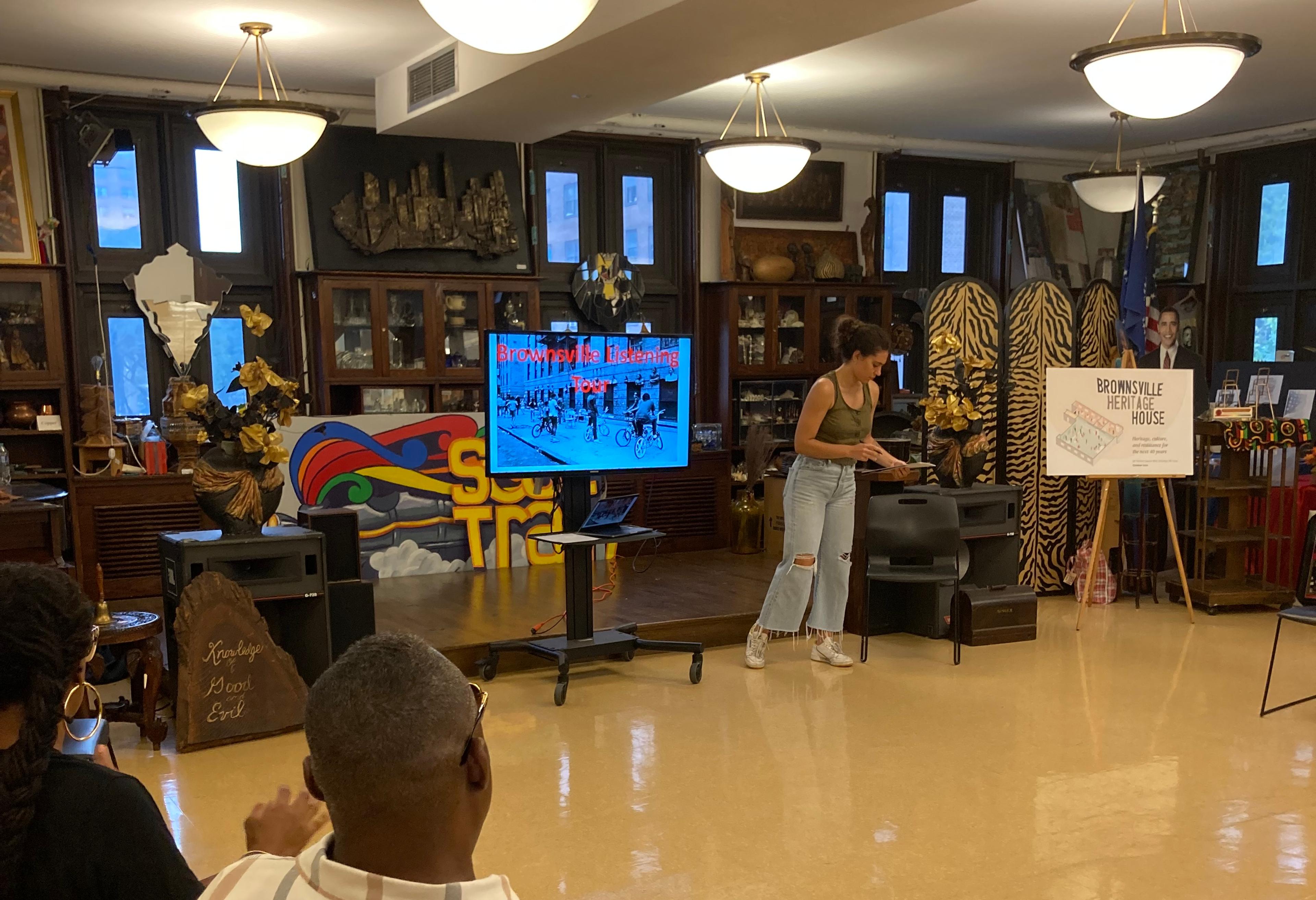 A young adult speaks to people seated in a circle beside a screen that reads "Brownsville Listening Tour"