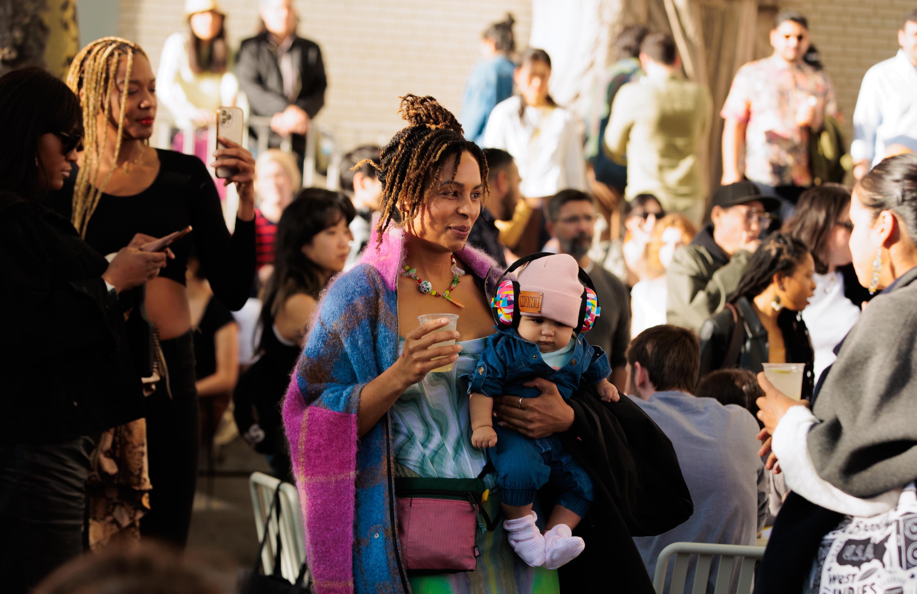 A person standing outside amongst other people, holds a drink and a baby with headphones on smiling