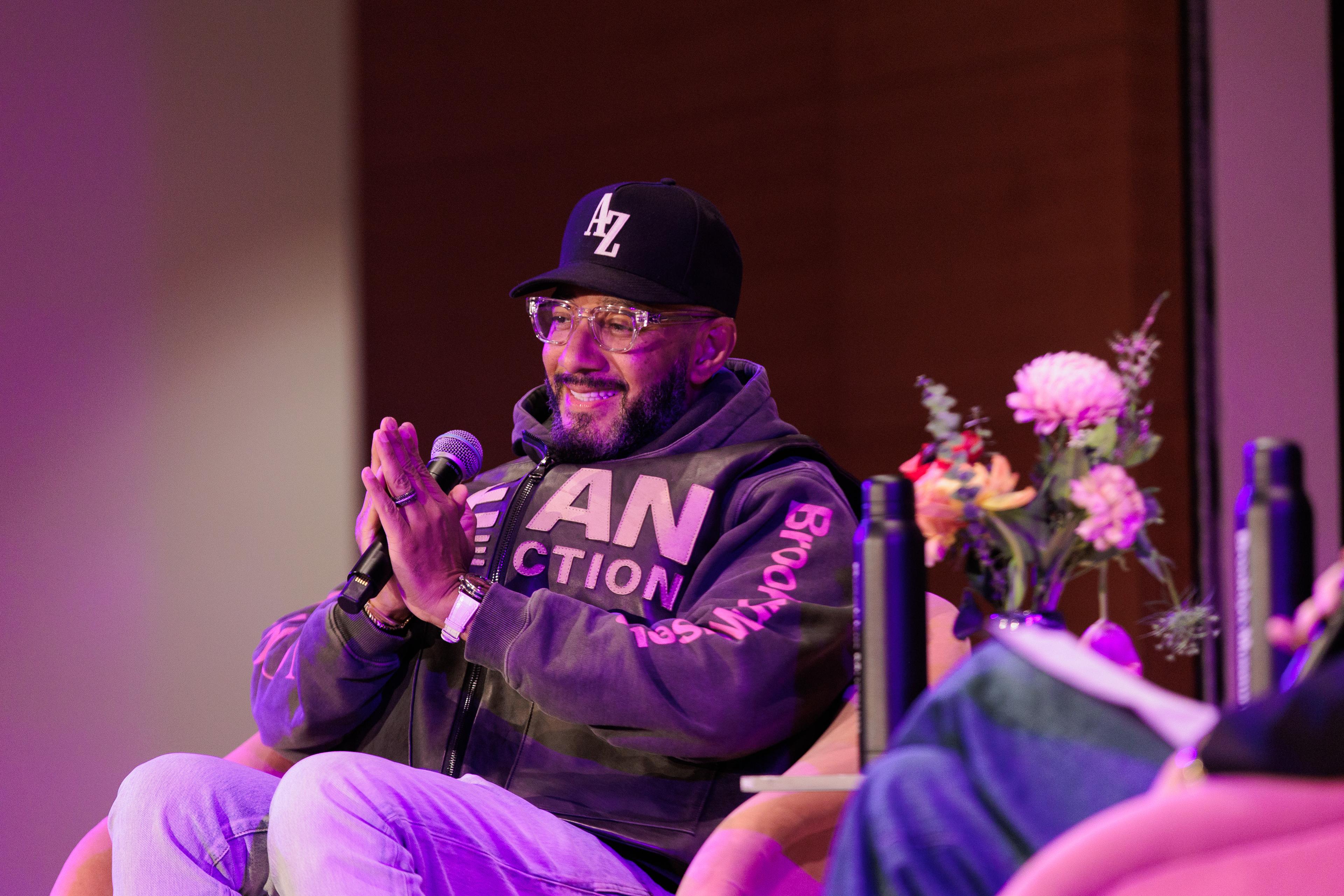 Smiling person holds a mic smiling while seated in a pink chair is wearing a sweat shirt that reads "Dean Collection" and "Brooklyn Museum"