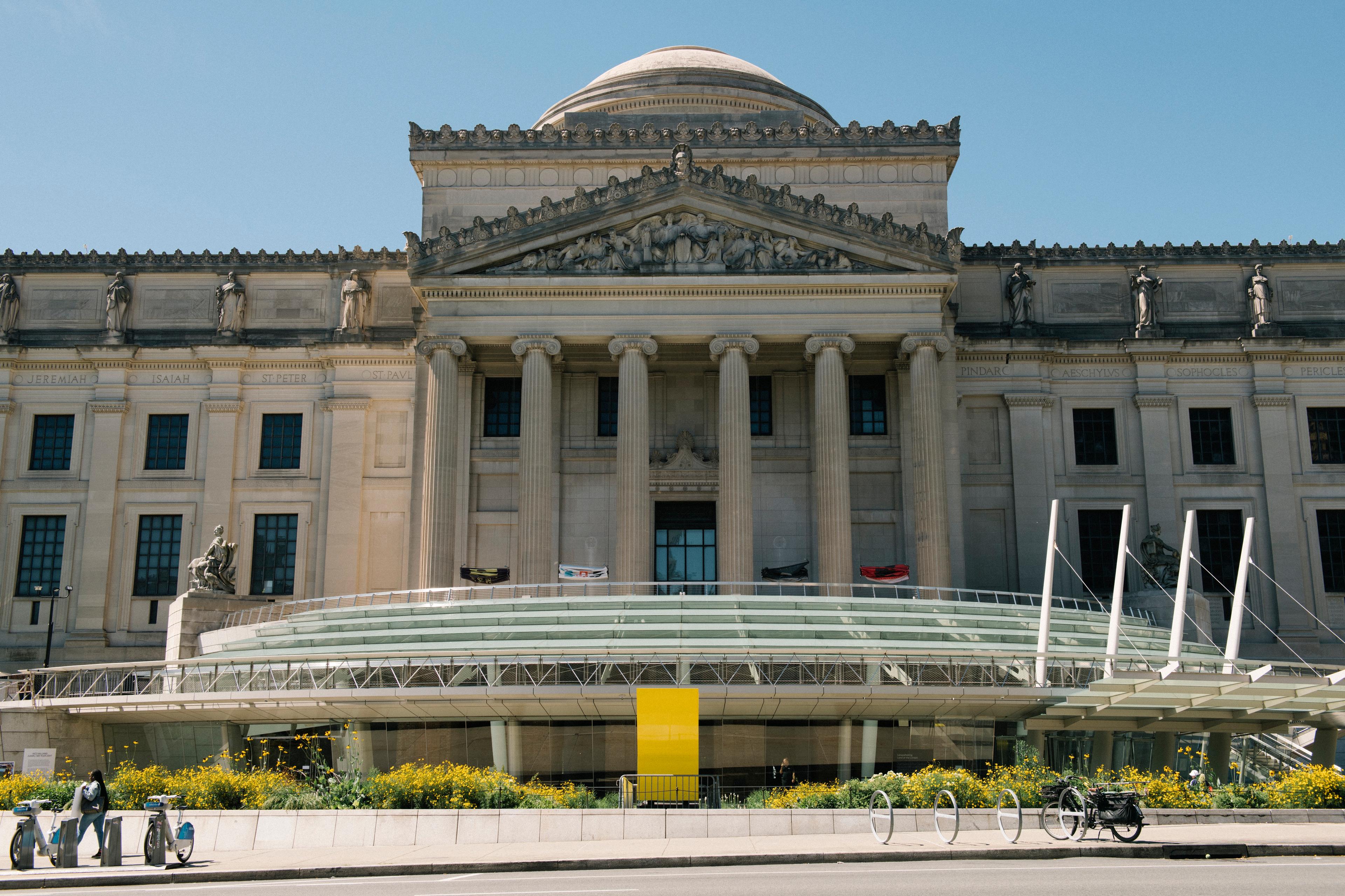 Large neoclassical building with pillars, dome roof, and glaza pavillion, with a landscaped plaza and bike racks in the foreground.