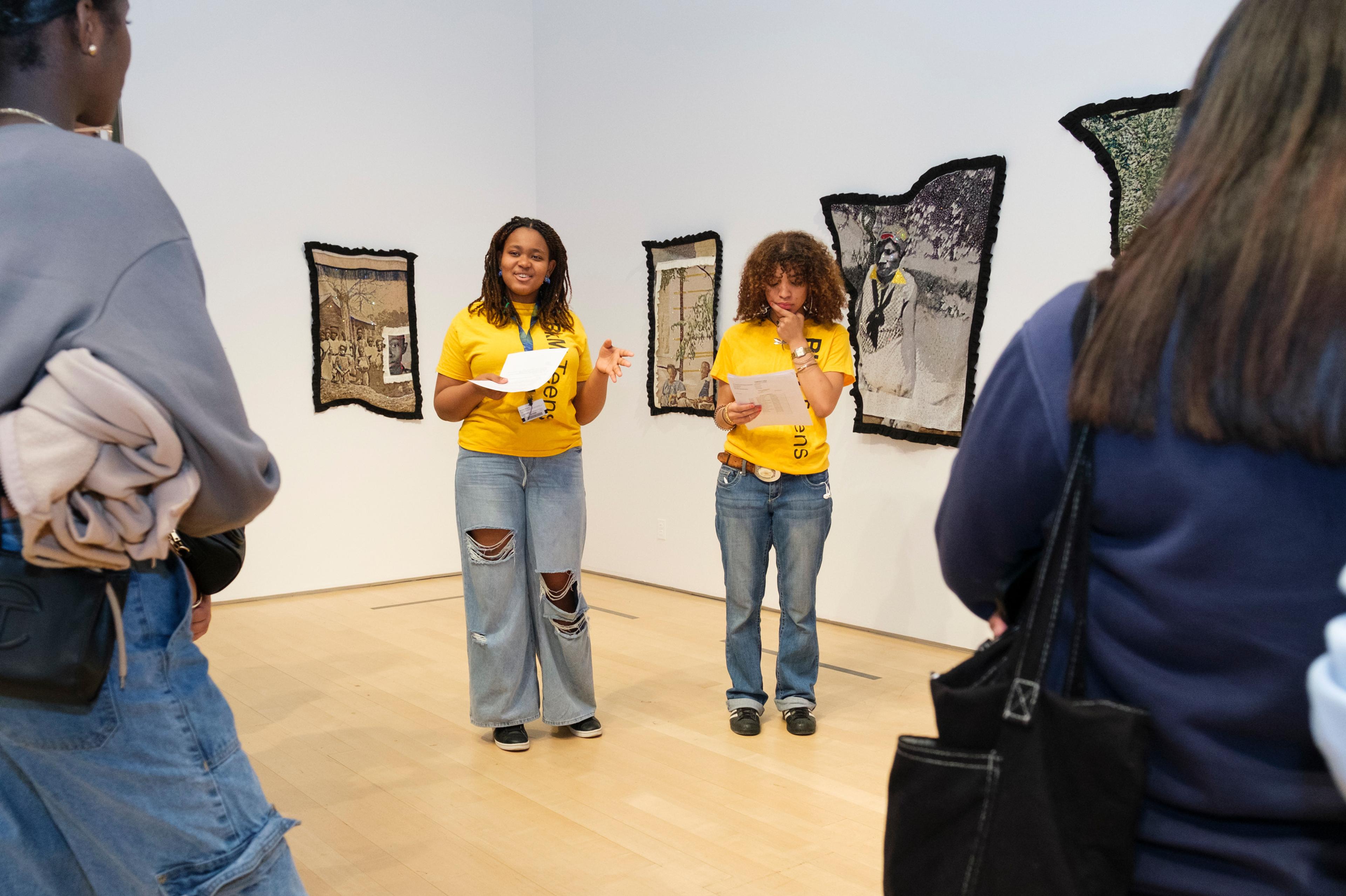 Two teenagers stand infront of hanging textile artworks, talking to a group of people