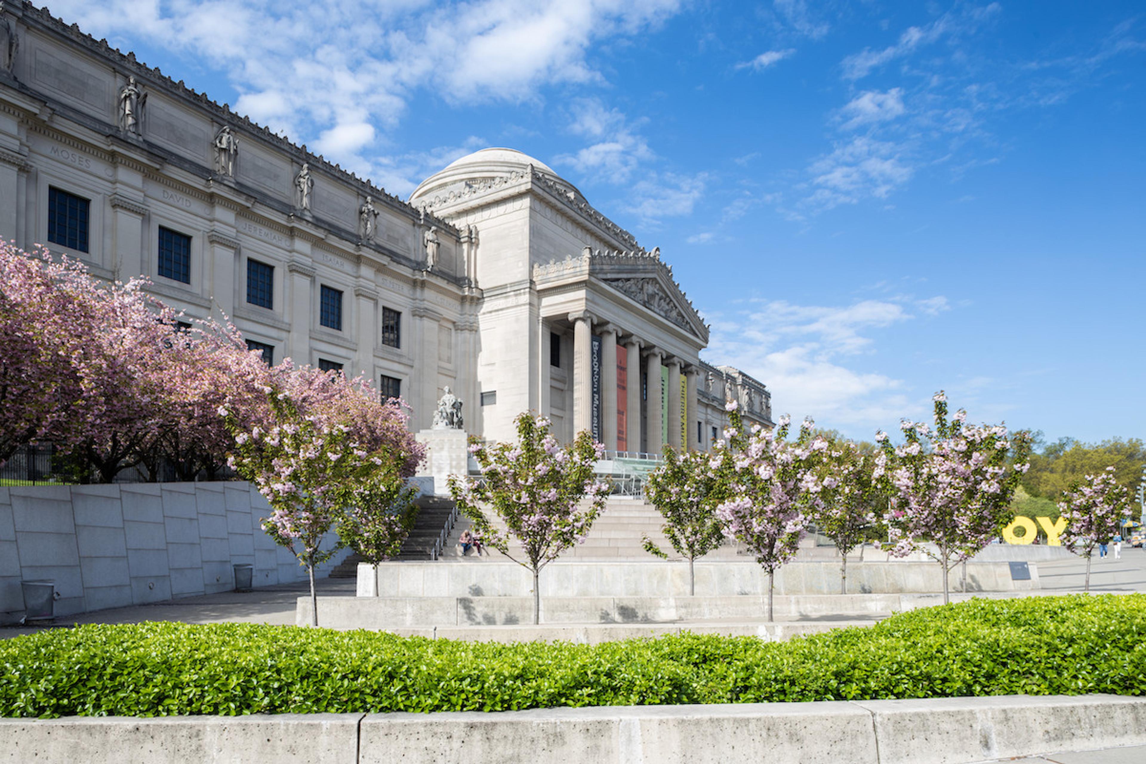 A photo of the Brooklyn Museum taken from one side of the plaza on a clear day, blooming trees are in the foreground