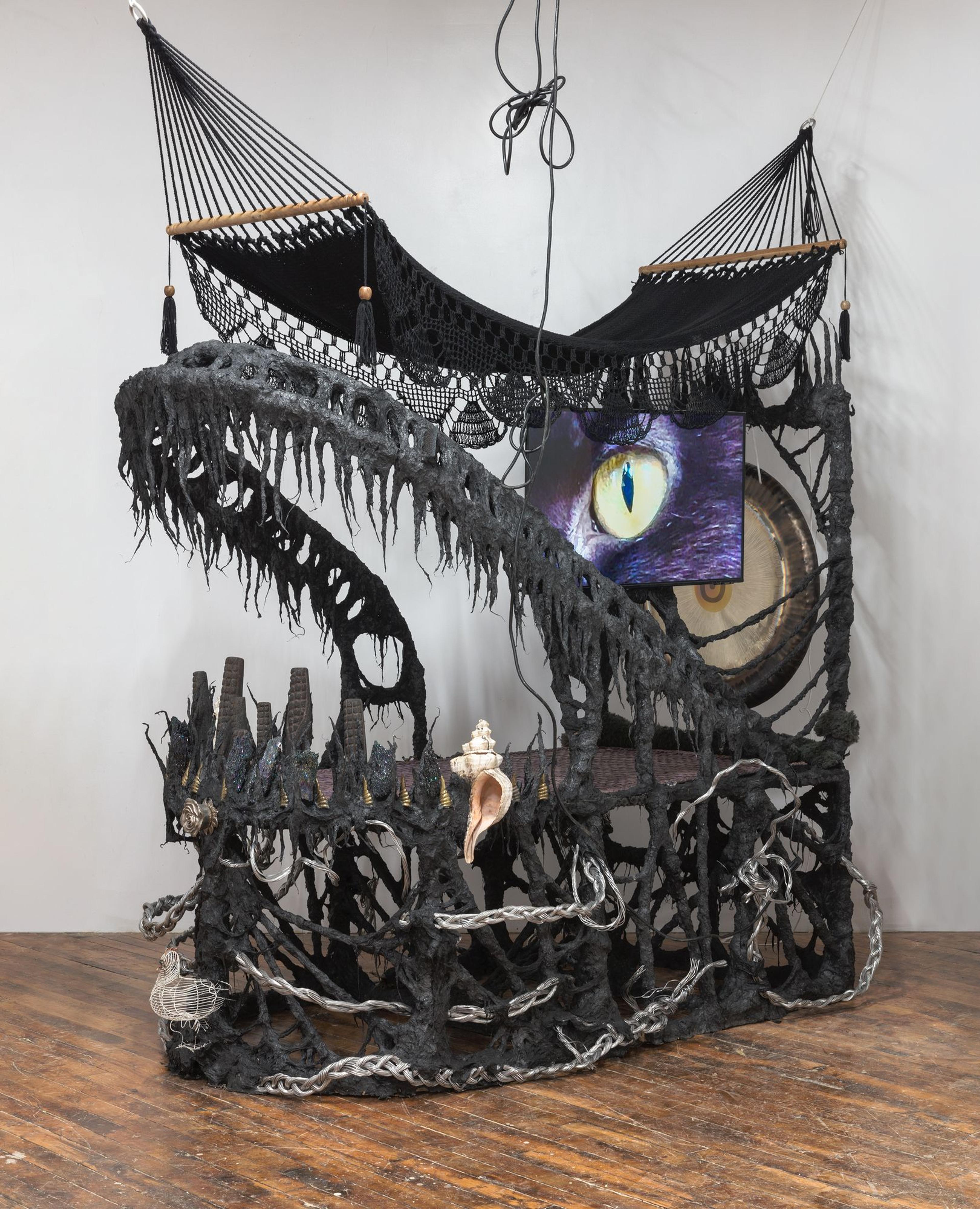 Guadalupe Maravilla (born El Salvador, 1976). Disease Thrower #0, 2022. Gong, hammock, LCD TV, ceremonial ash, pyrite crystals, volcanic rock, steel, wood, cotton and glue mixture, plastic, loofah, objects collected from a ritual of retracing the artist's original migration route, 118 × 123 × 64 in. (299.7 × 312.4 × 162.6 cm). Courtesy of the artist and P·P·O·W, New York. © Guadalupe Maravilla. (Photo: Stan Narten)