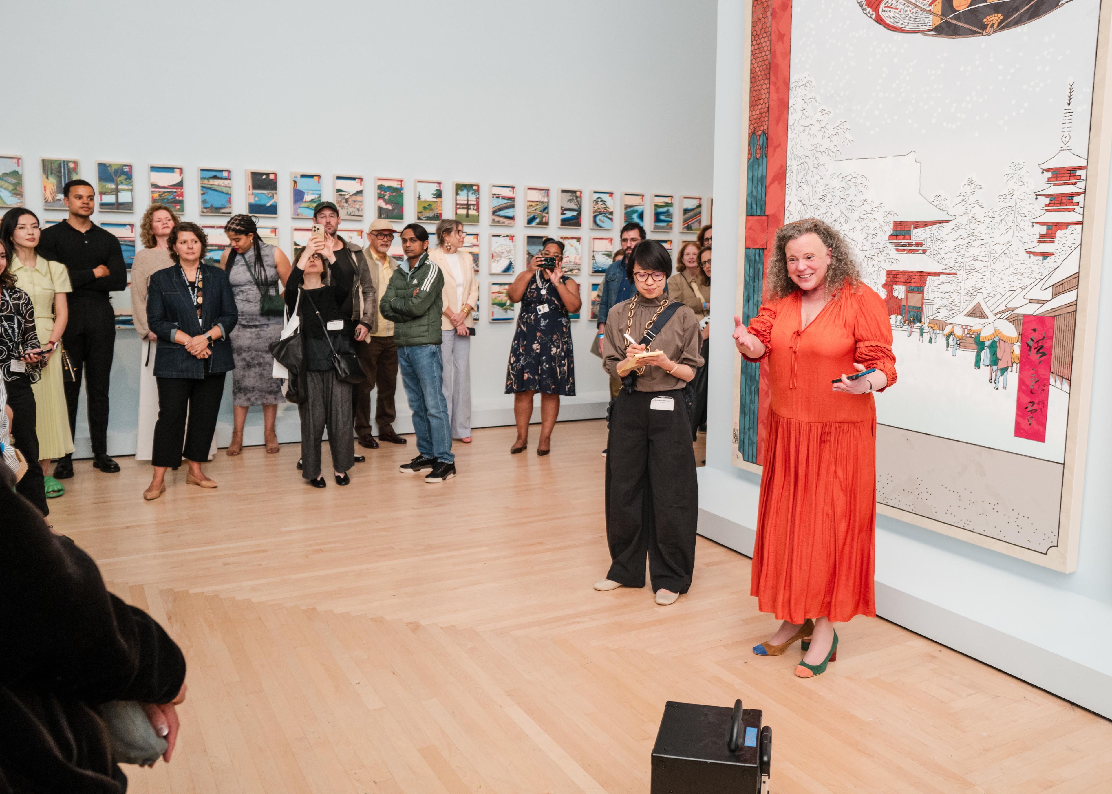 A person in an orange dress smiles and gestures while talking to a small audience in an art gallery