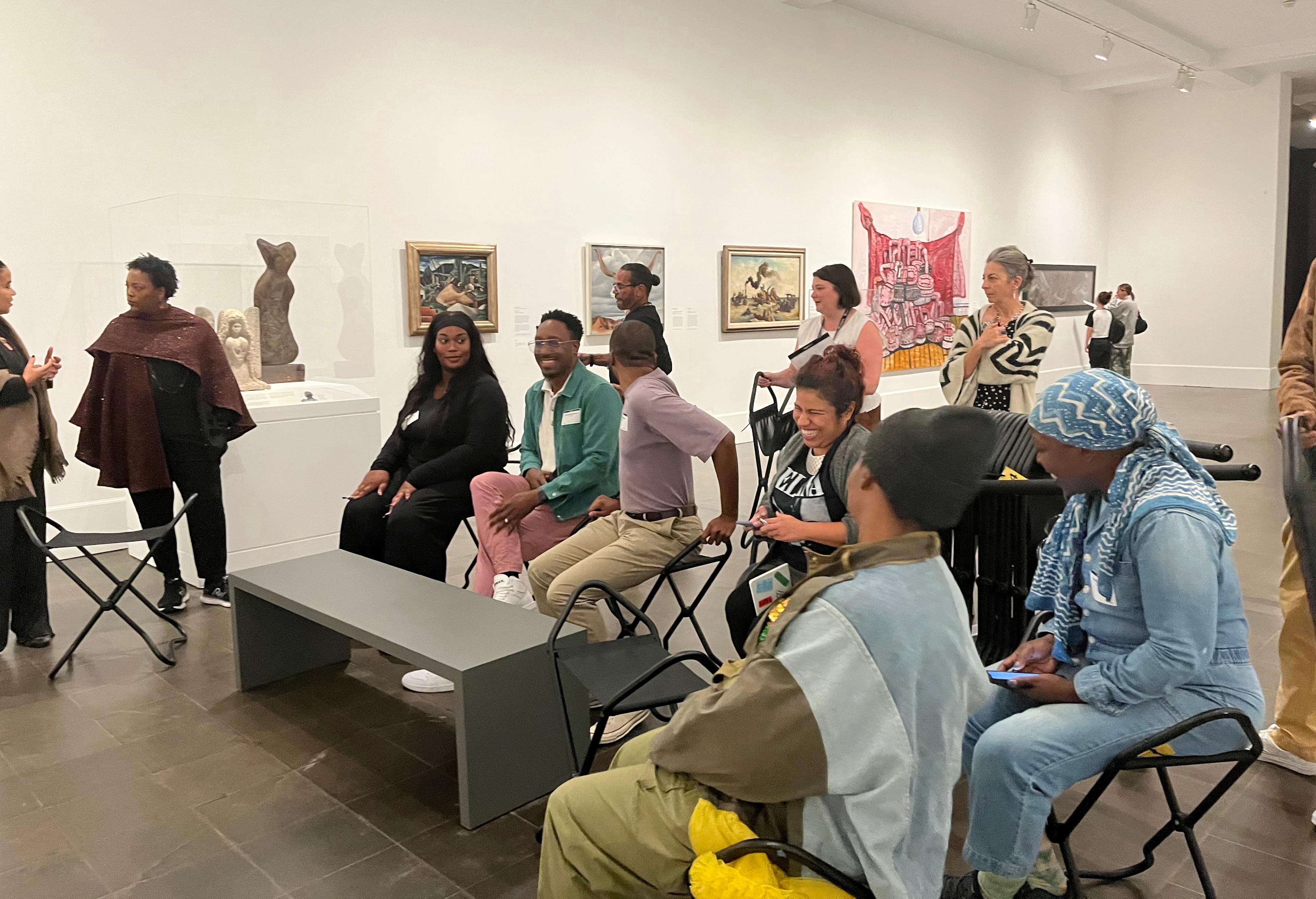 Size adults seated in folding stools smile and laugh with one another in an art gallery with other adults standing near by