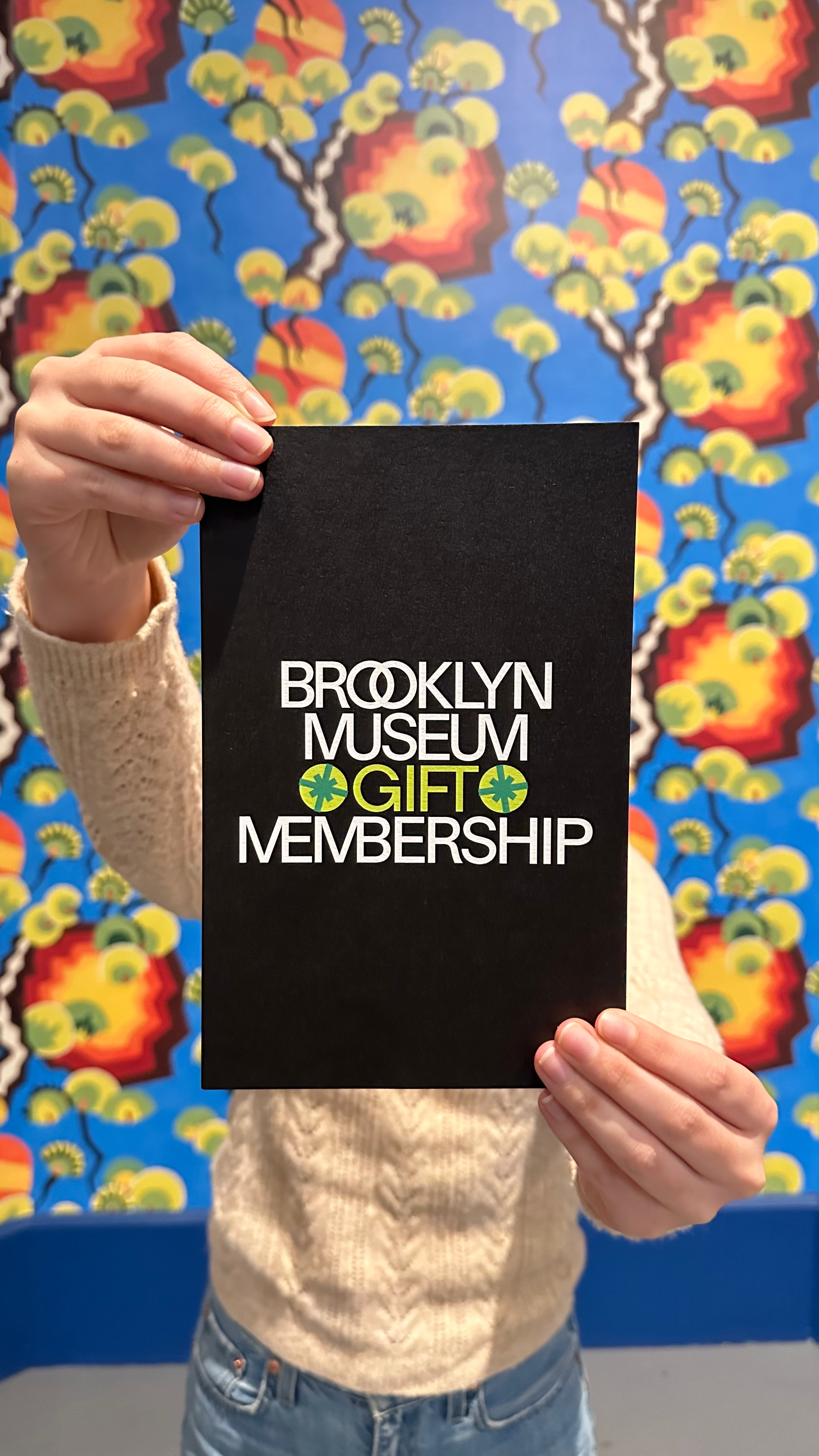 Person holding up a black card reading “Brooklyn Museum Gift Membership” in front of a colorful wall