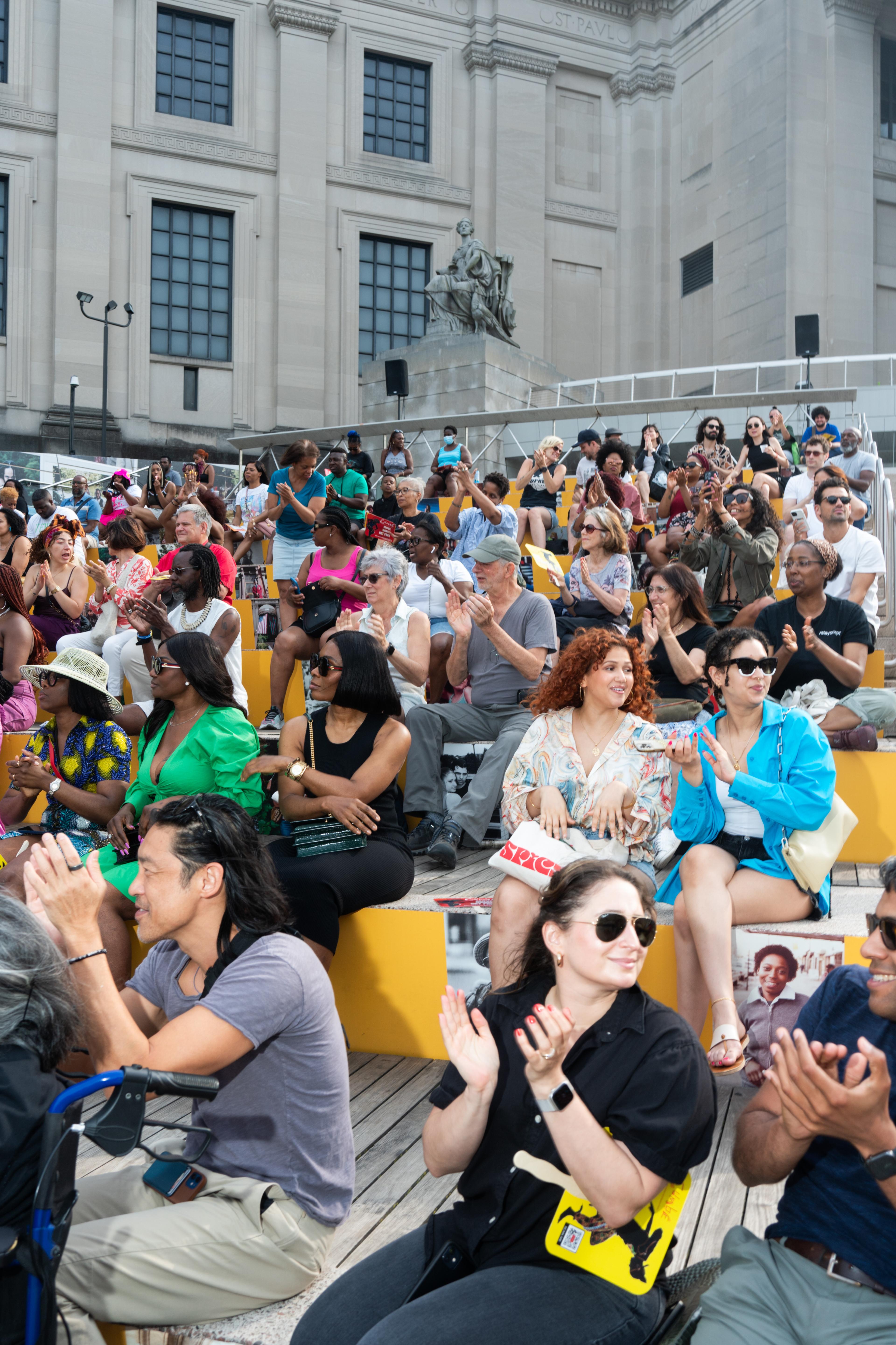 Dozens of adults sit on outdoor steps clapping, smiling and looking around