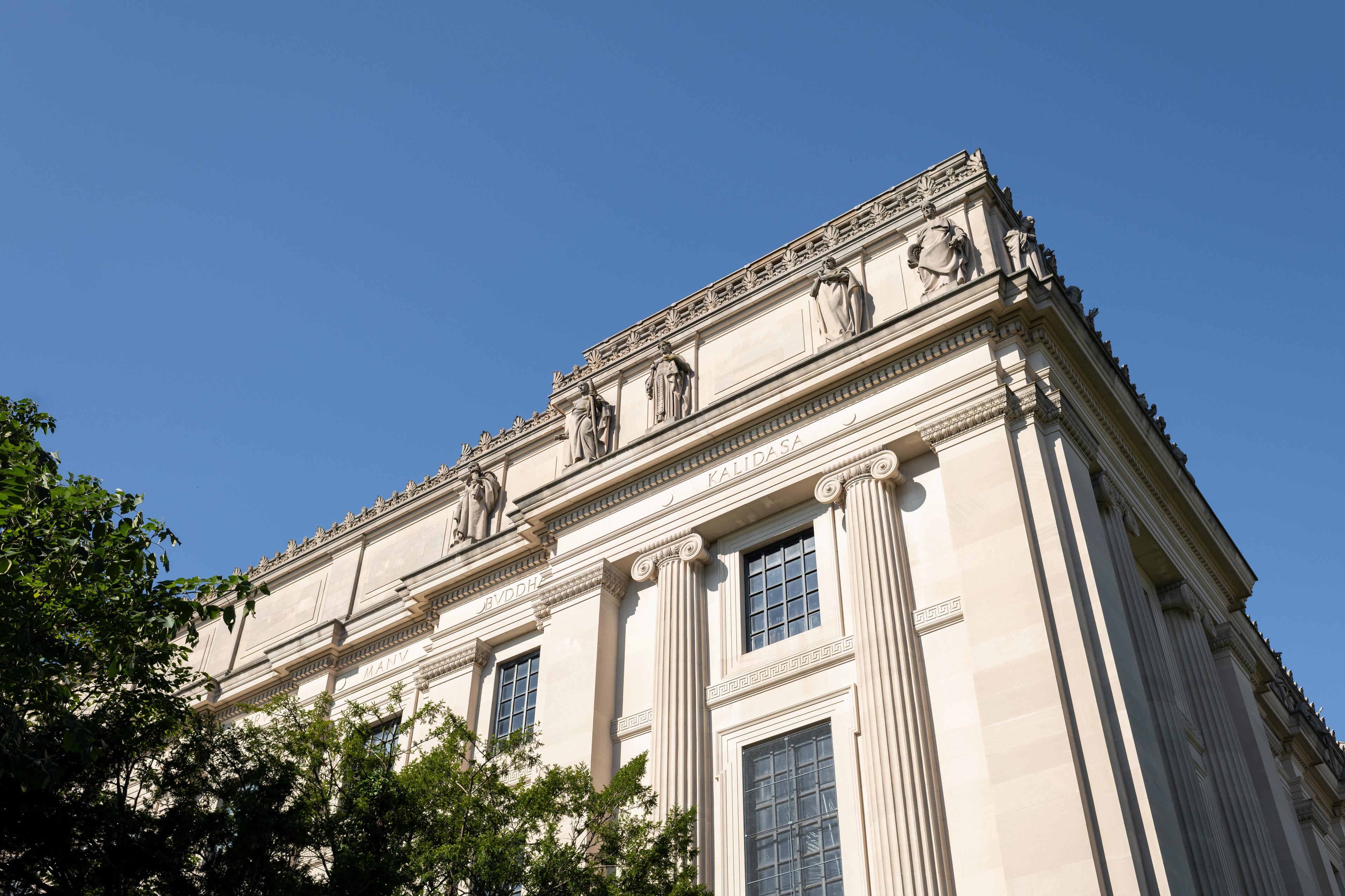 Corner of a large building featuring columns and windows in the mid section and statues along the top