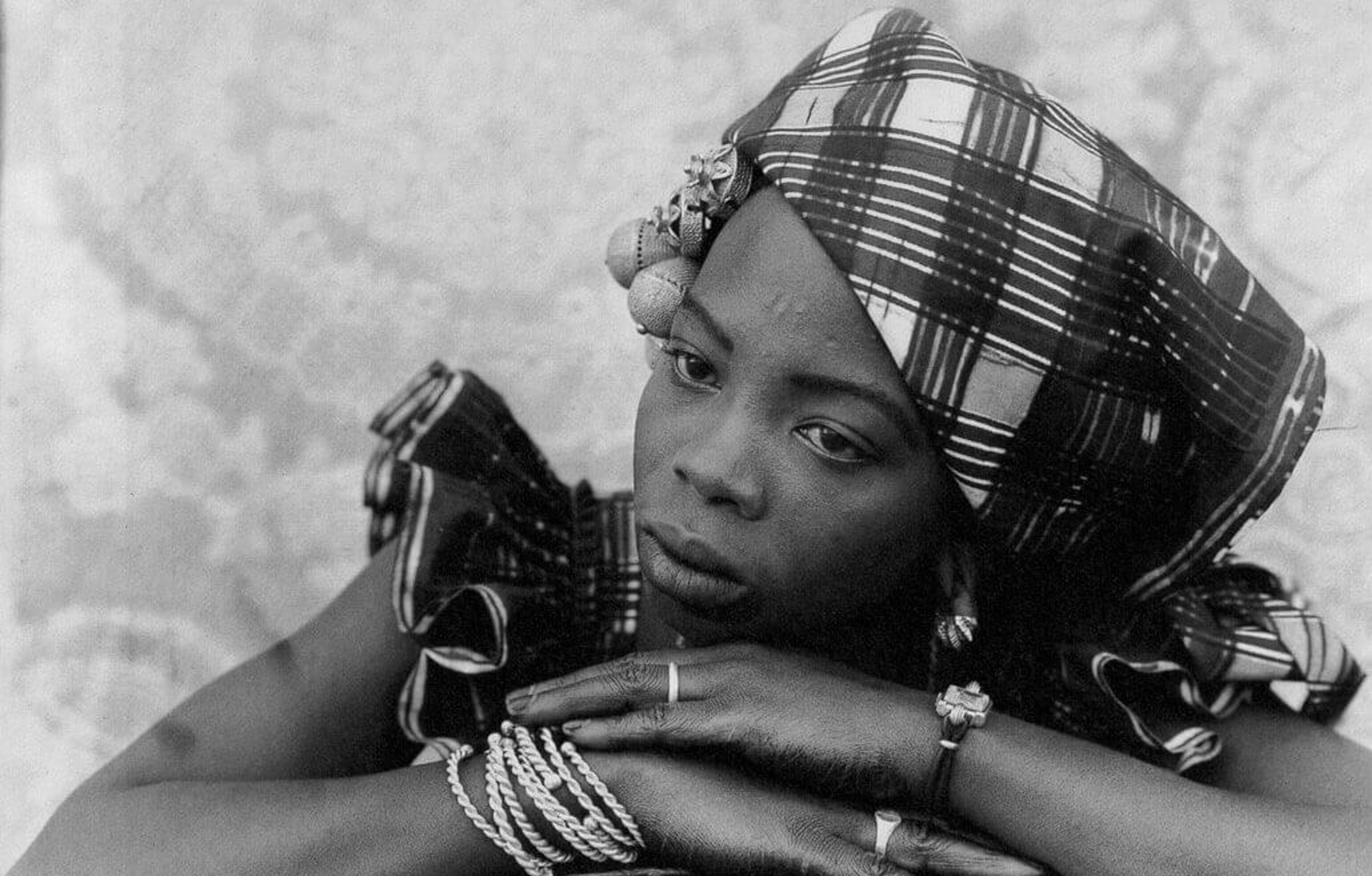 Black-and-white photograph of a woman resting her chin on crossed arms, wearing plaid headwrap and jewelry