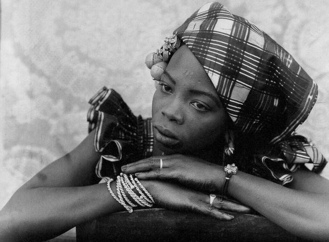 Black-and-white photograph of a woman resting her chin on crossed arms, wearing plaid headwrap and jewelry