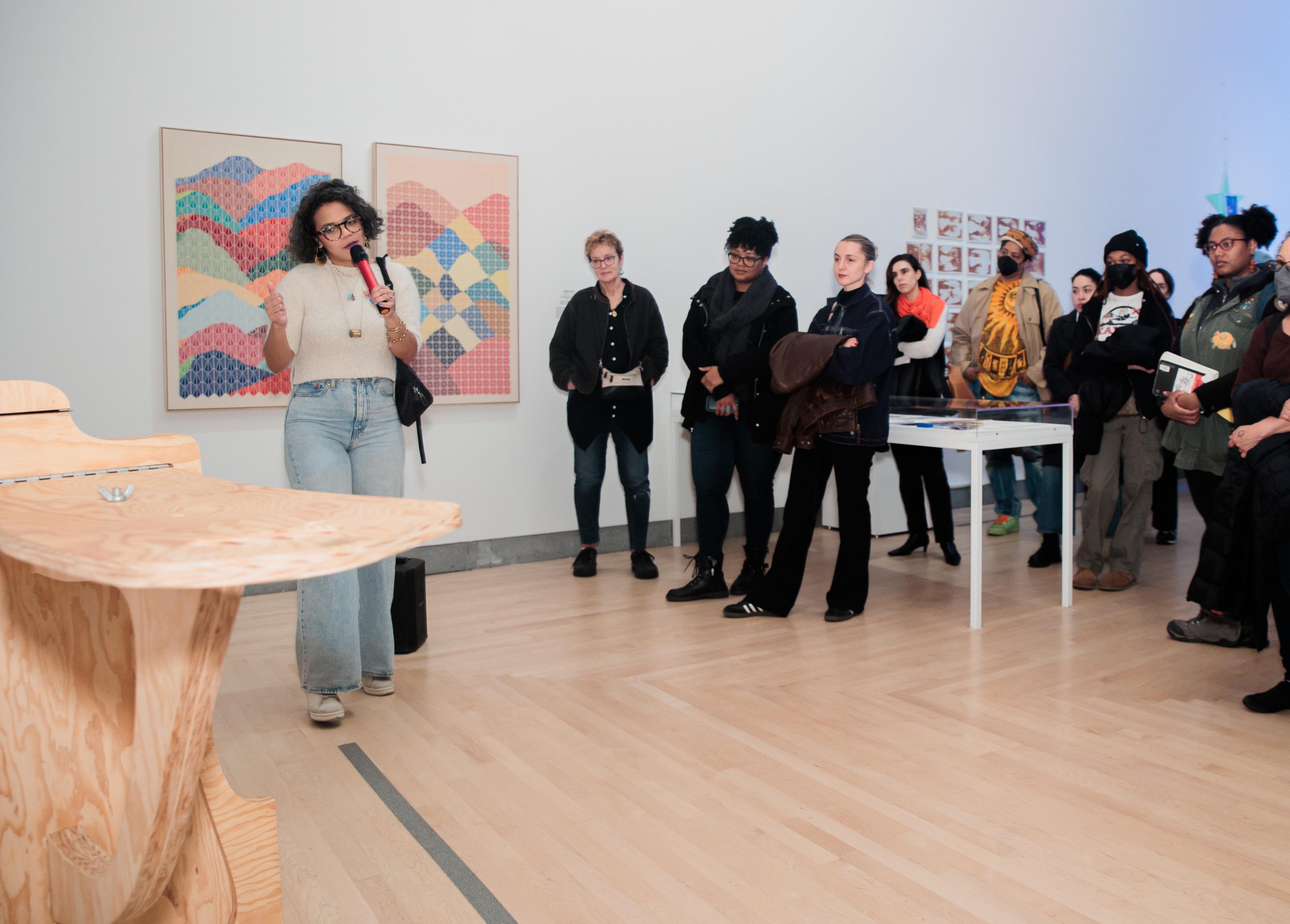 In a white art gallery a person holding a mic gestures towards a wooden artwork while speaking to a crowd of vistors