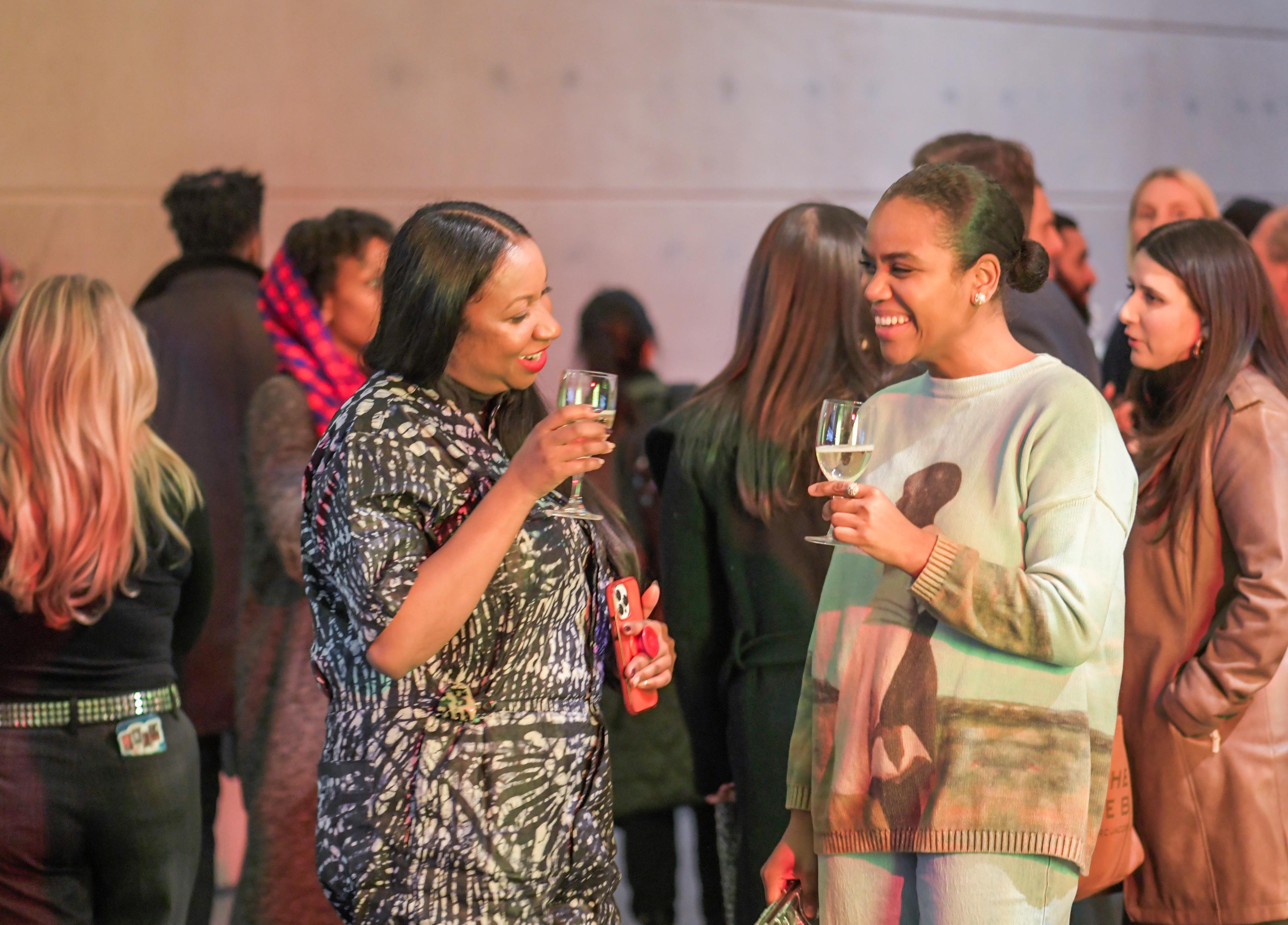 Two visitors holding drinks are talking and laughing amongst a crowd of people