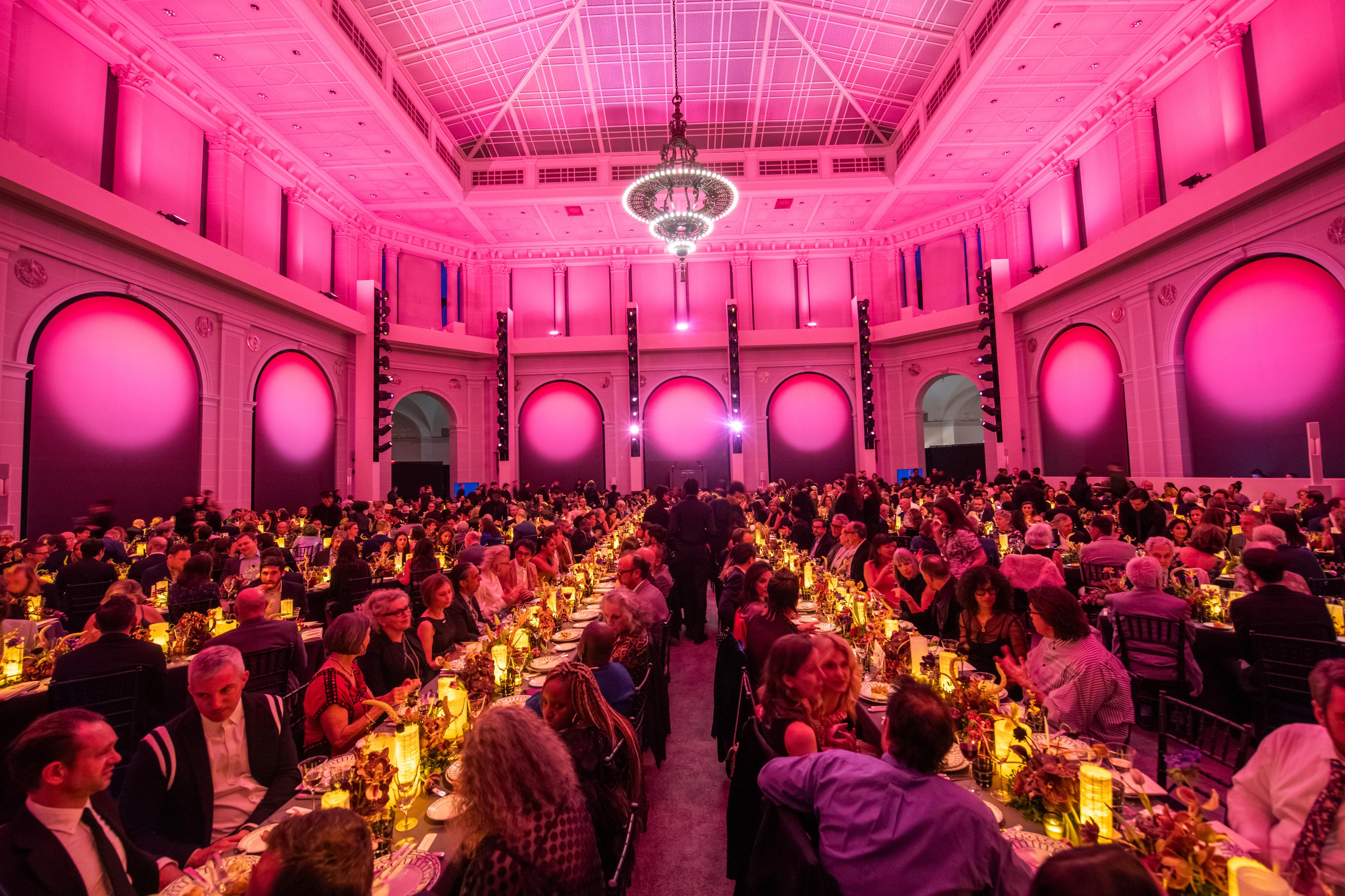 A large room glows in pink and gold lighting as tables of diners sit beneath a large chandelier