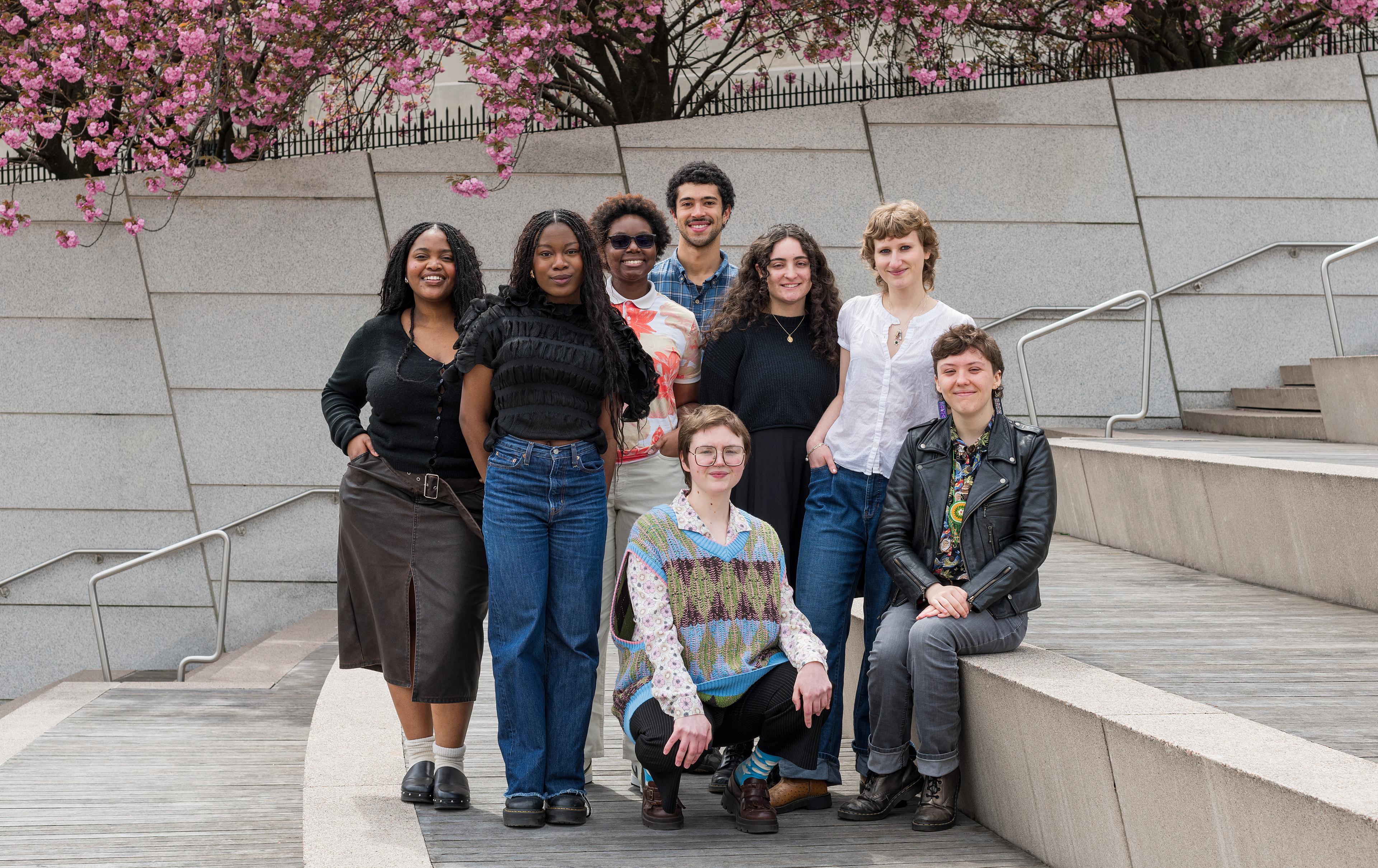 Group of eight young adults pose together smiling on stairs with cherry blossoms in the background