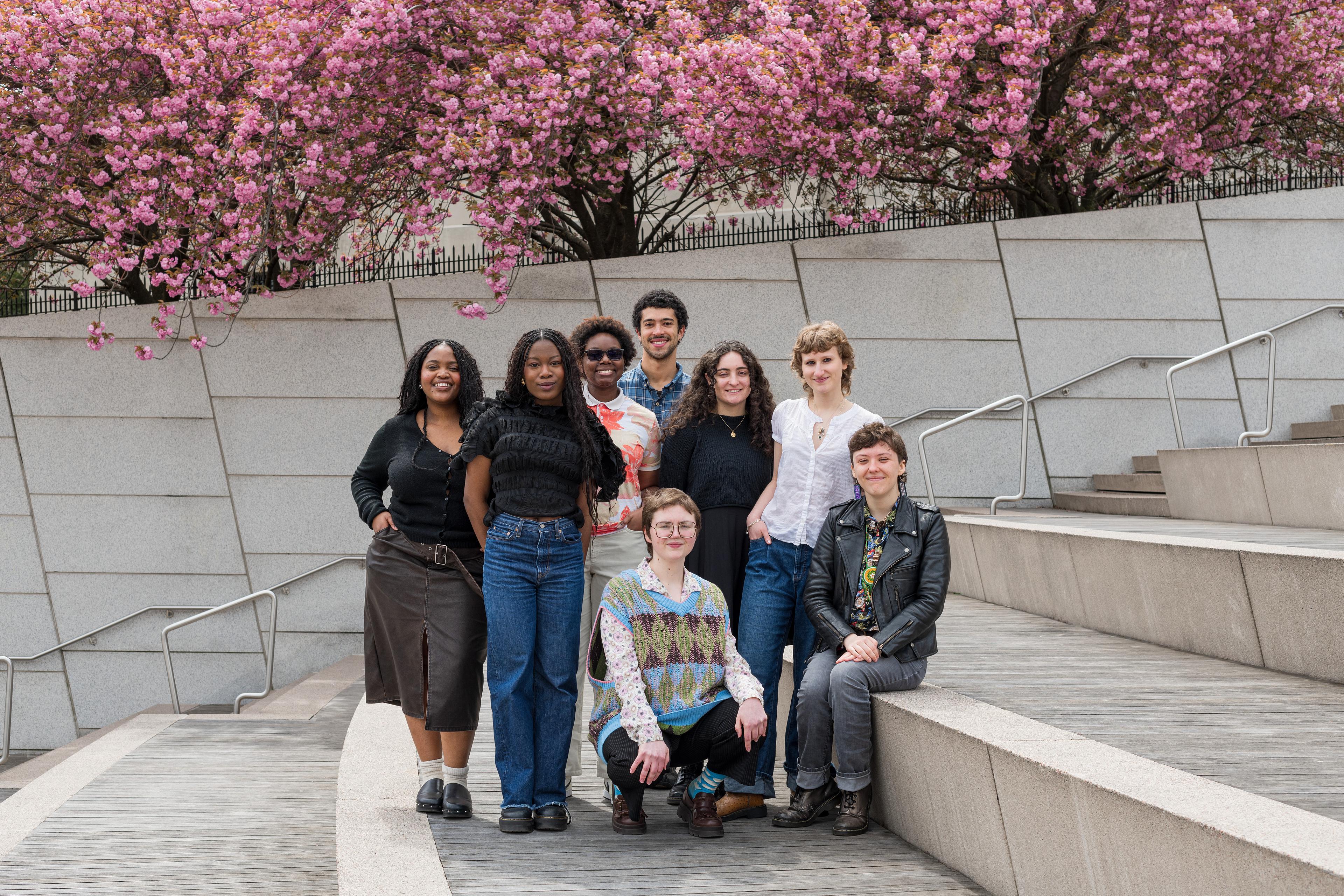 Group of eight young adults pose together smiling on stairs with cherry blossoms in the background