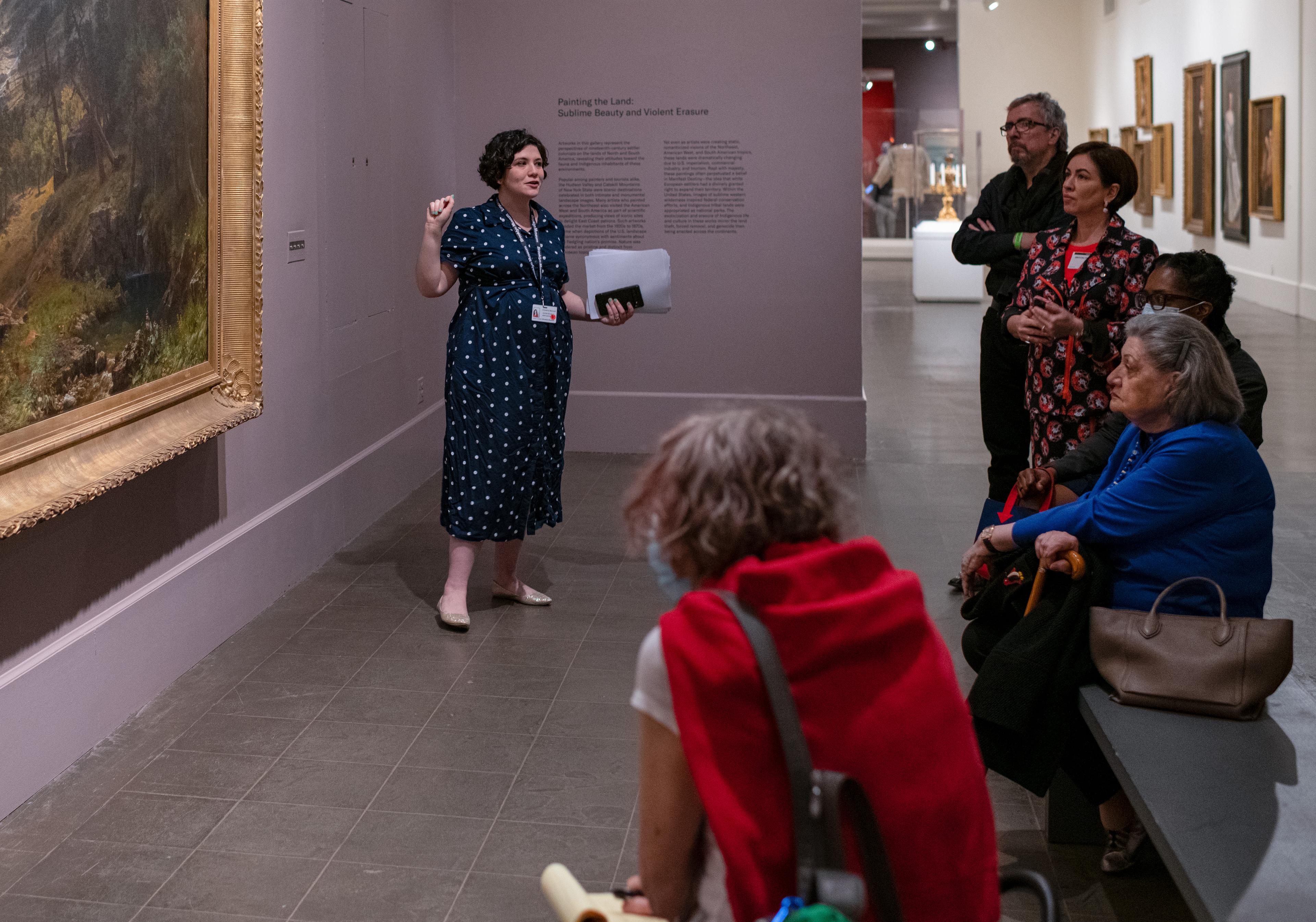 An educator stands in front of a large landscape painting, speaking to a group of five people