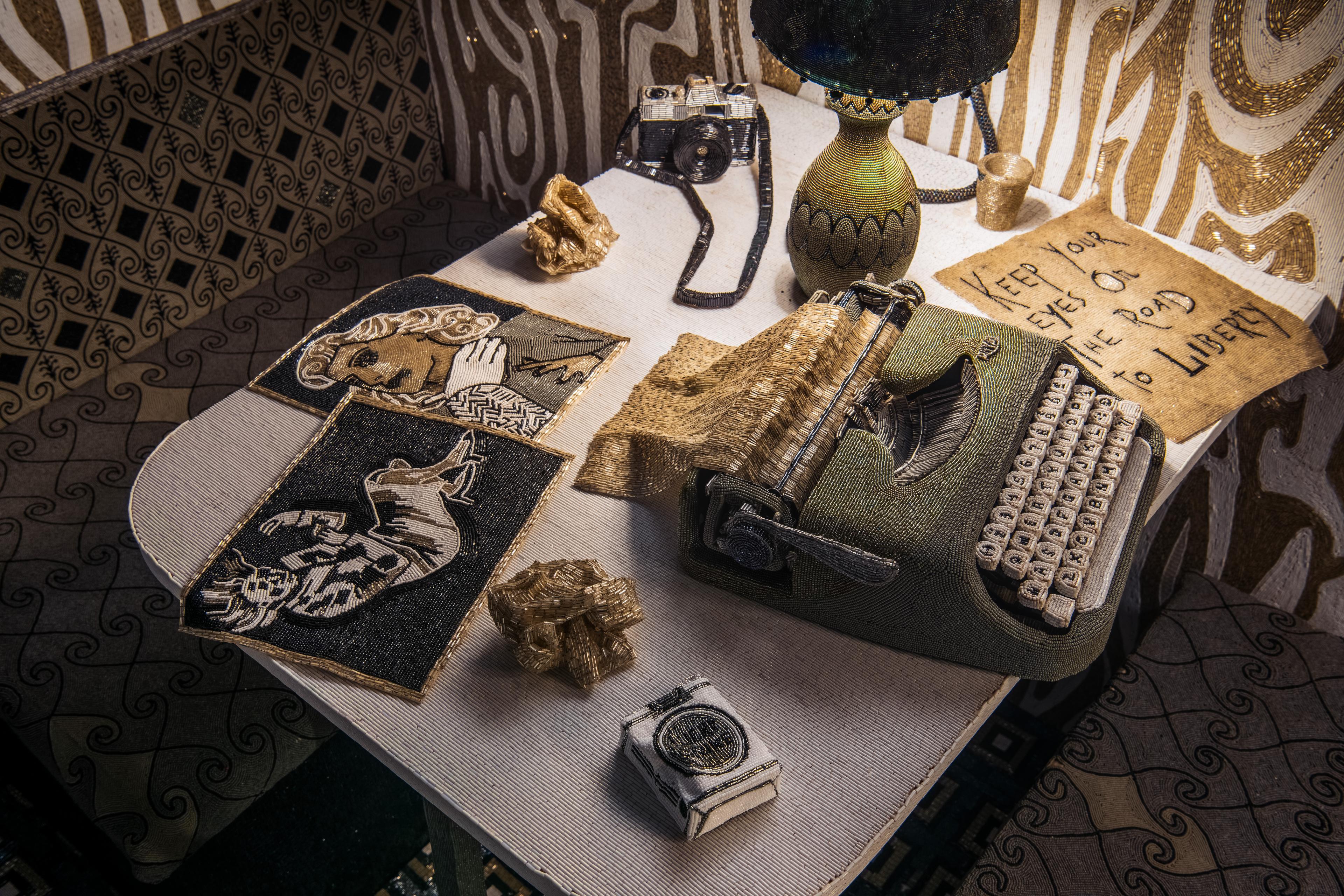 Interior detail of a table with bench seats. Table has a typewriter, photos, camera, lamp, pack of cigarettes. Everything is made of beads.