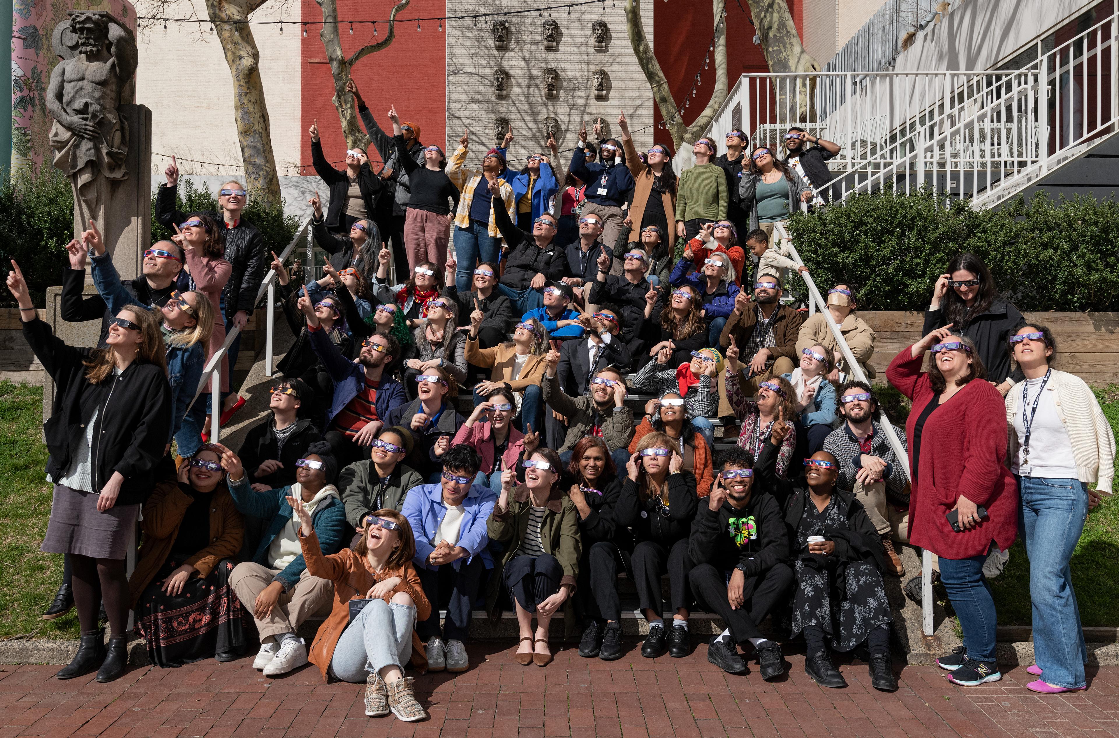 Dozens of adults sit outside on steps wearing glasses and facing and pointing at the sun