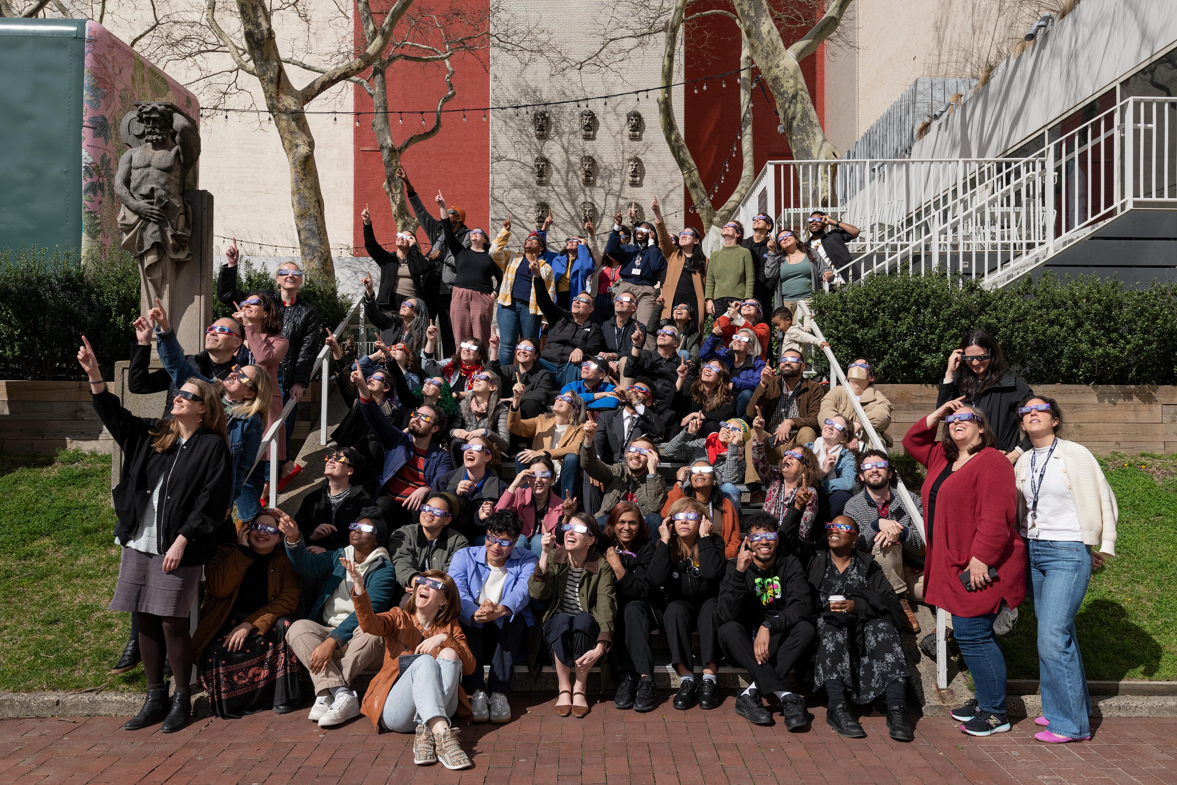 Dozens of adults sit outside on steps wearing glasses and facing and pointing at the sun