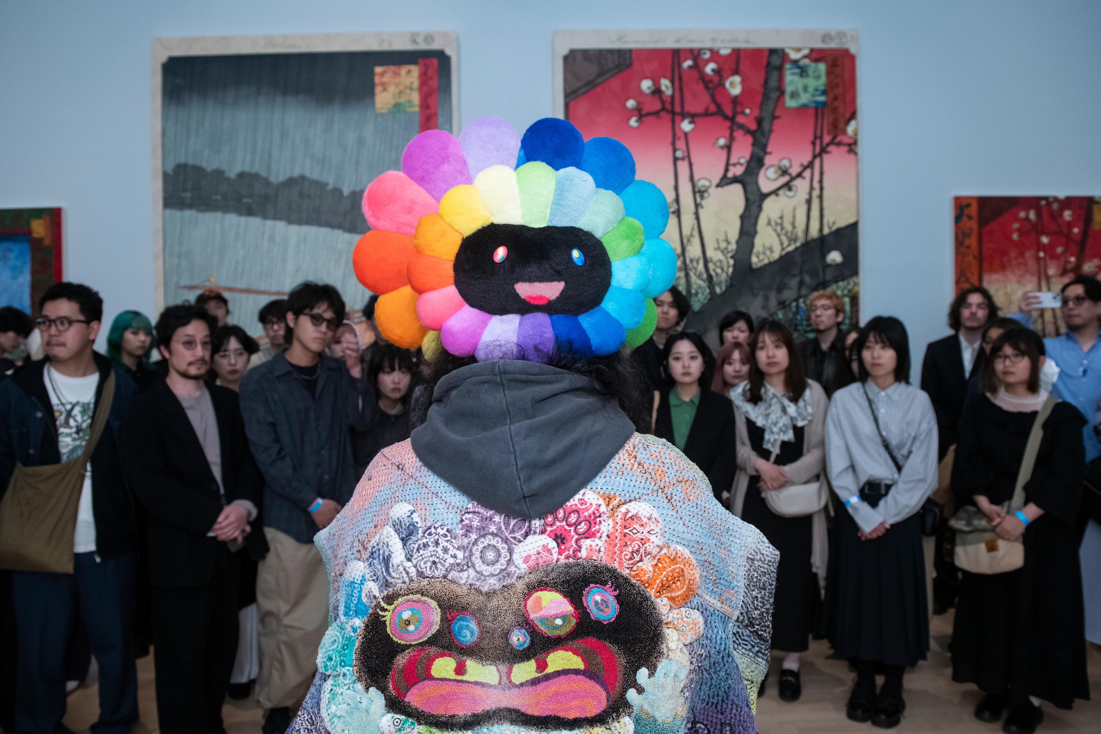 Artist (Takashi Murakami) wears a multi-colored flower hat and jacket and speaking to a crowd of people in a gallery