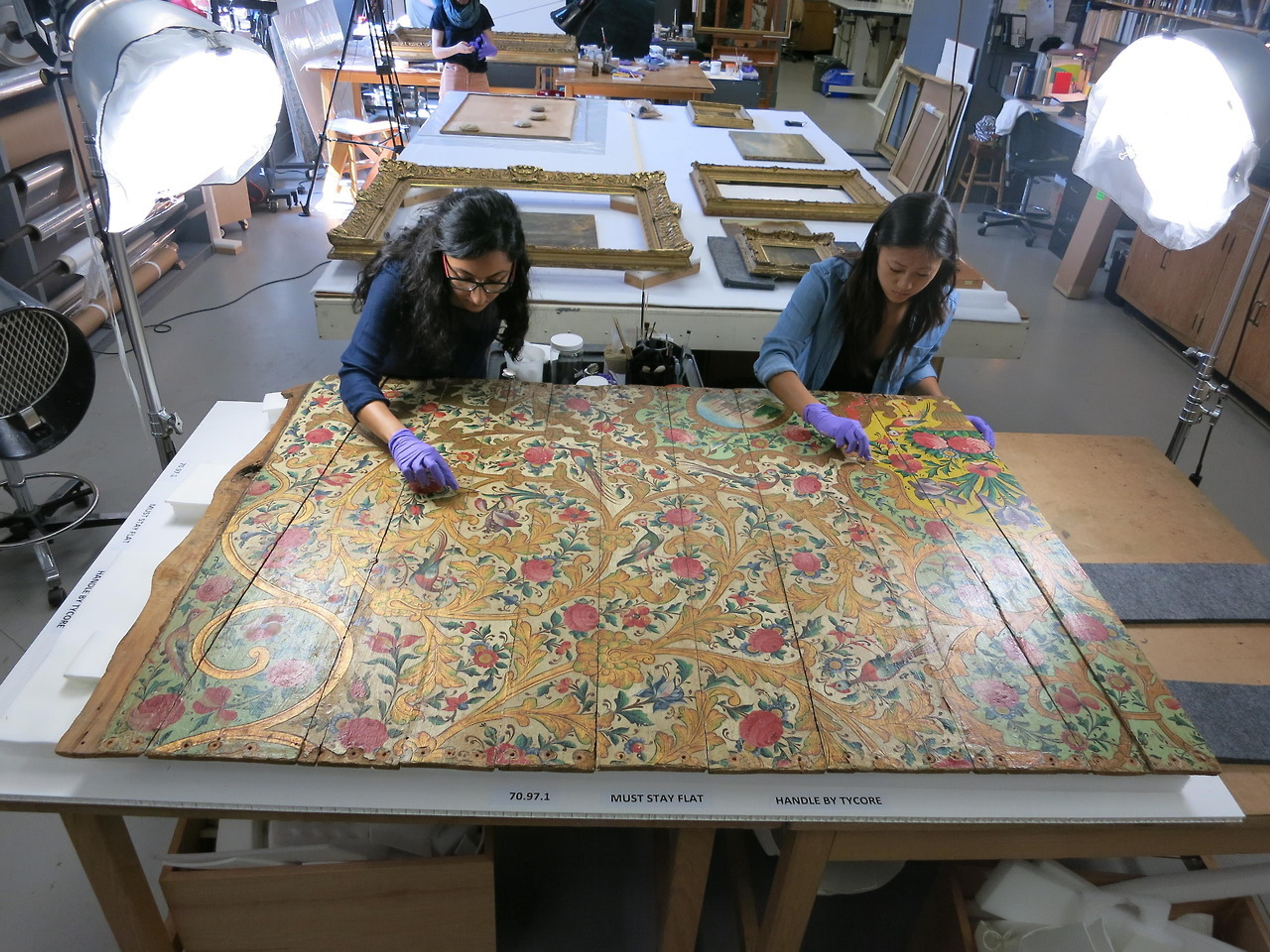 Two staff members wearing gloves work on a large, painted, wooden panel in the conservation laboratory