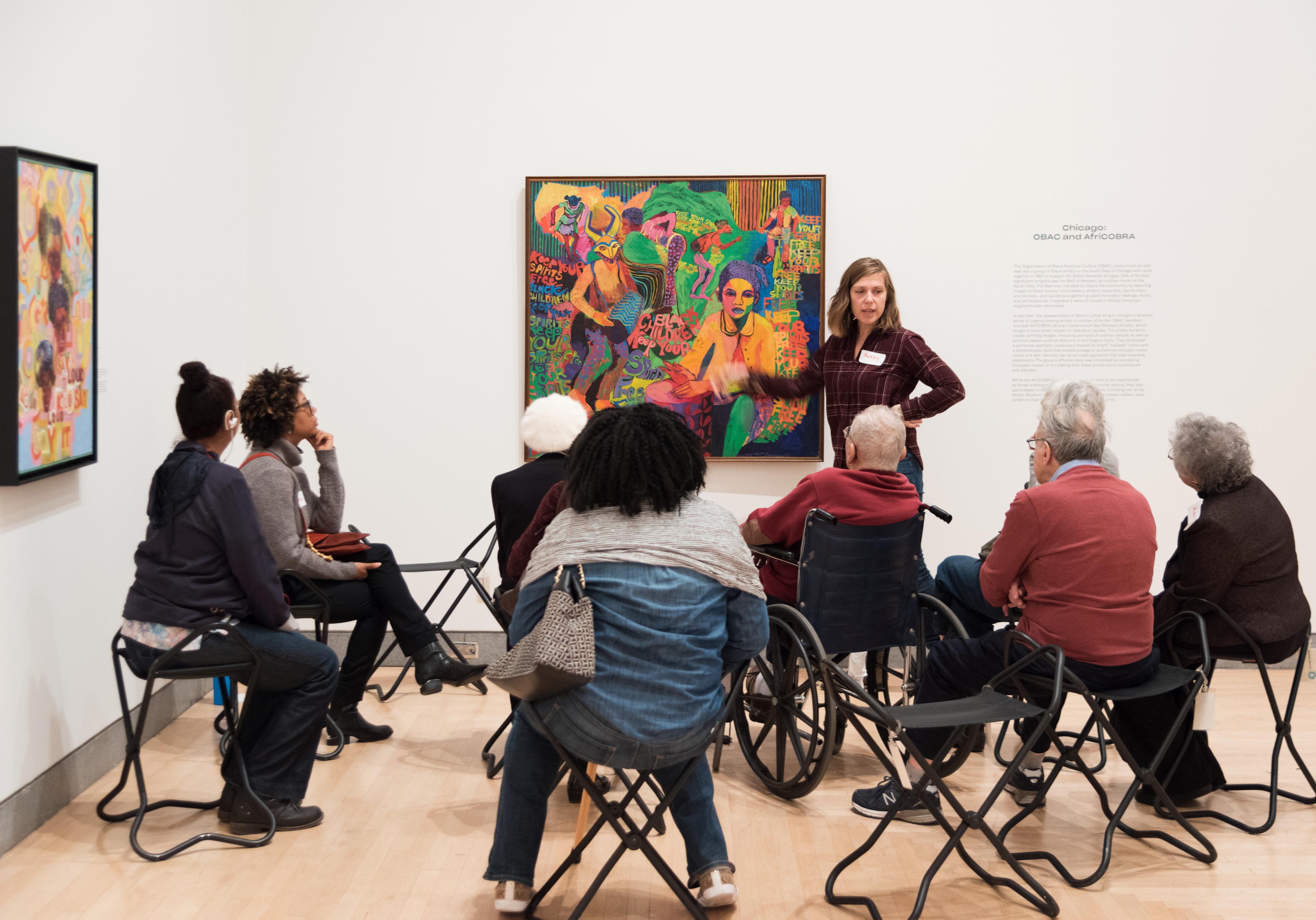 A group of adults in folding stools and a wheel chair circle around an artwork with a standing instructor speaking to them