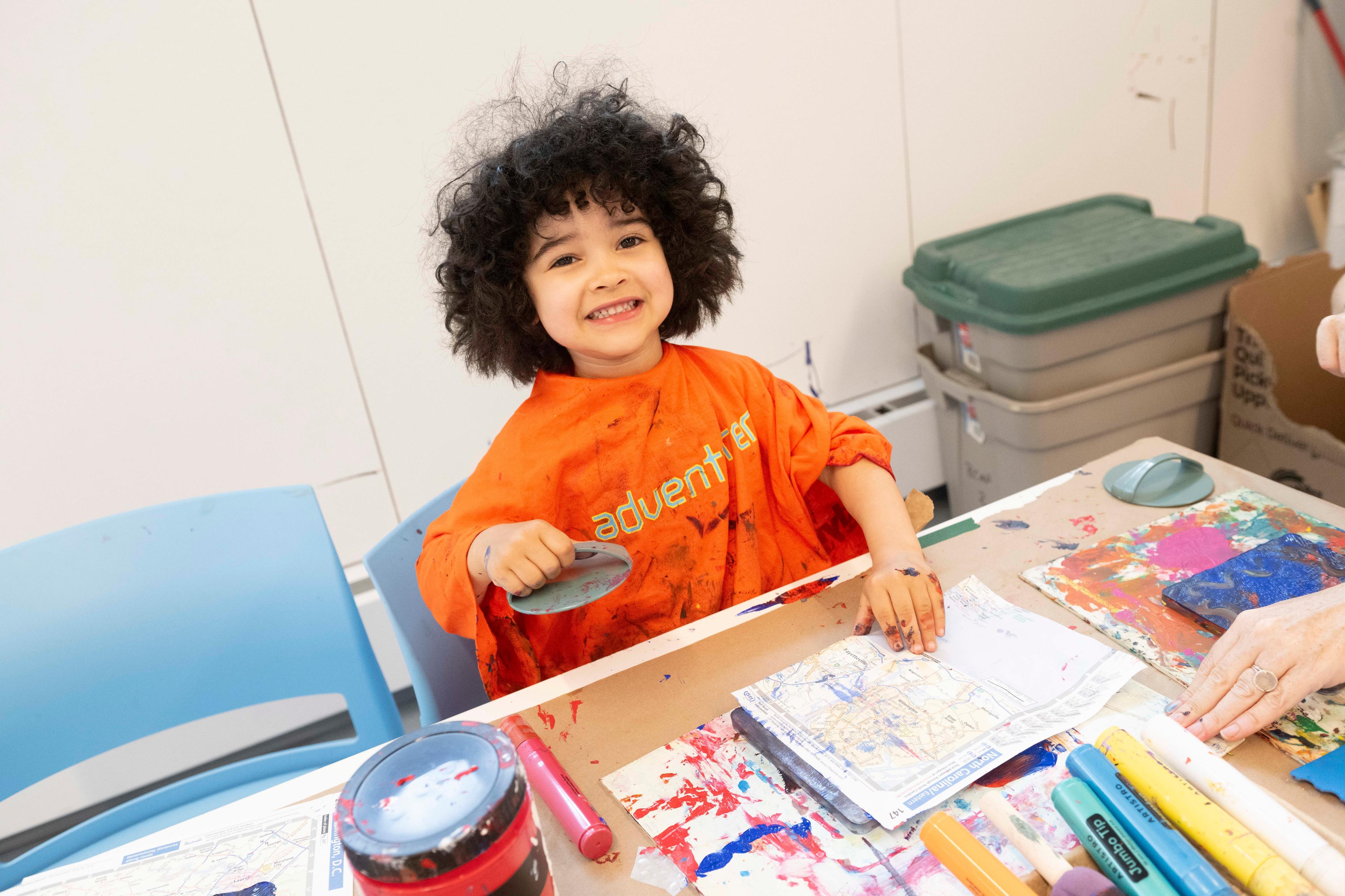 Child wearing an orange shirt that reads "adventurer" smiles with art materials