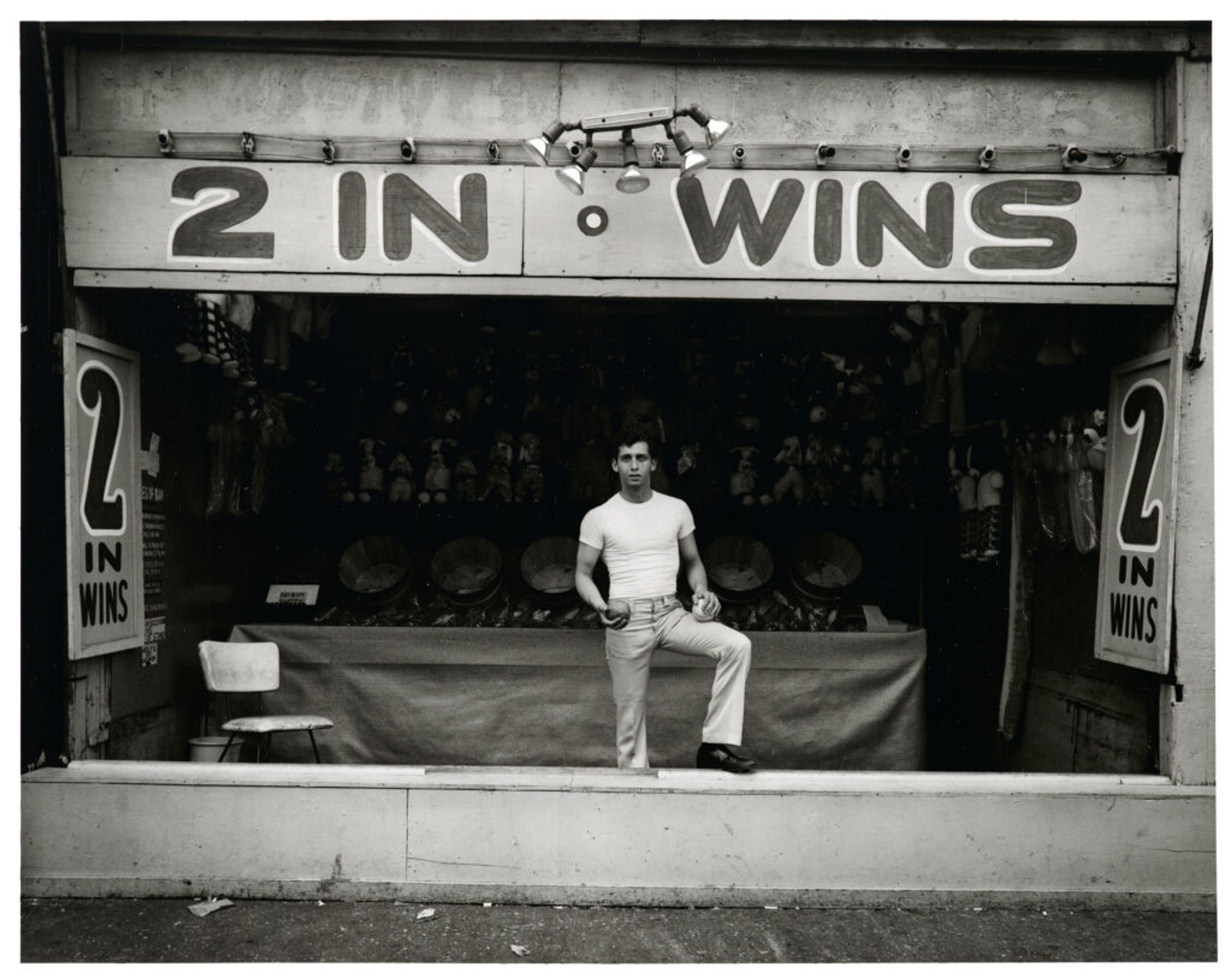 Man standing in carnival game booth labeled “2 in Wins”