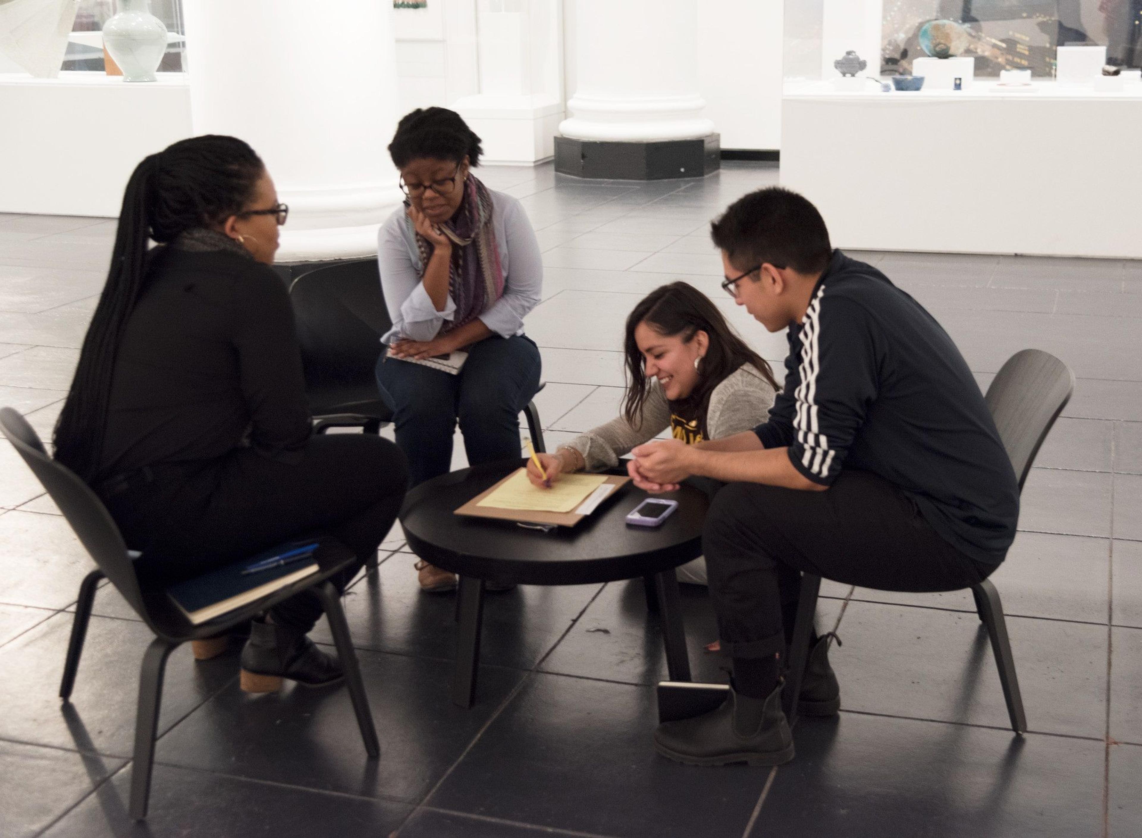 Four adults sit around a table, three look on as the fourth writes on paper