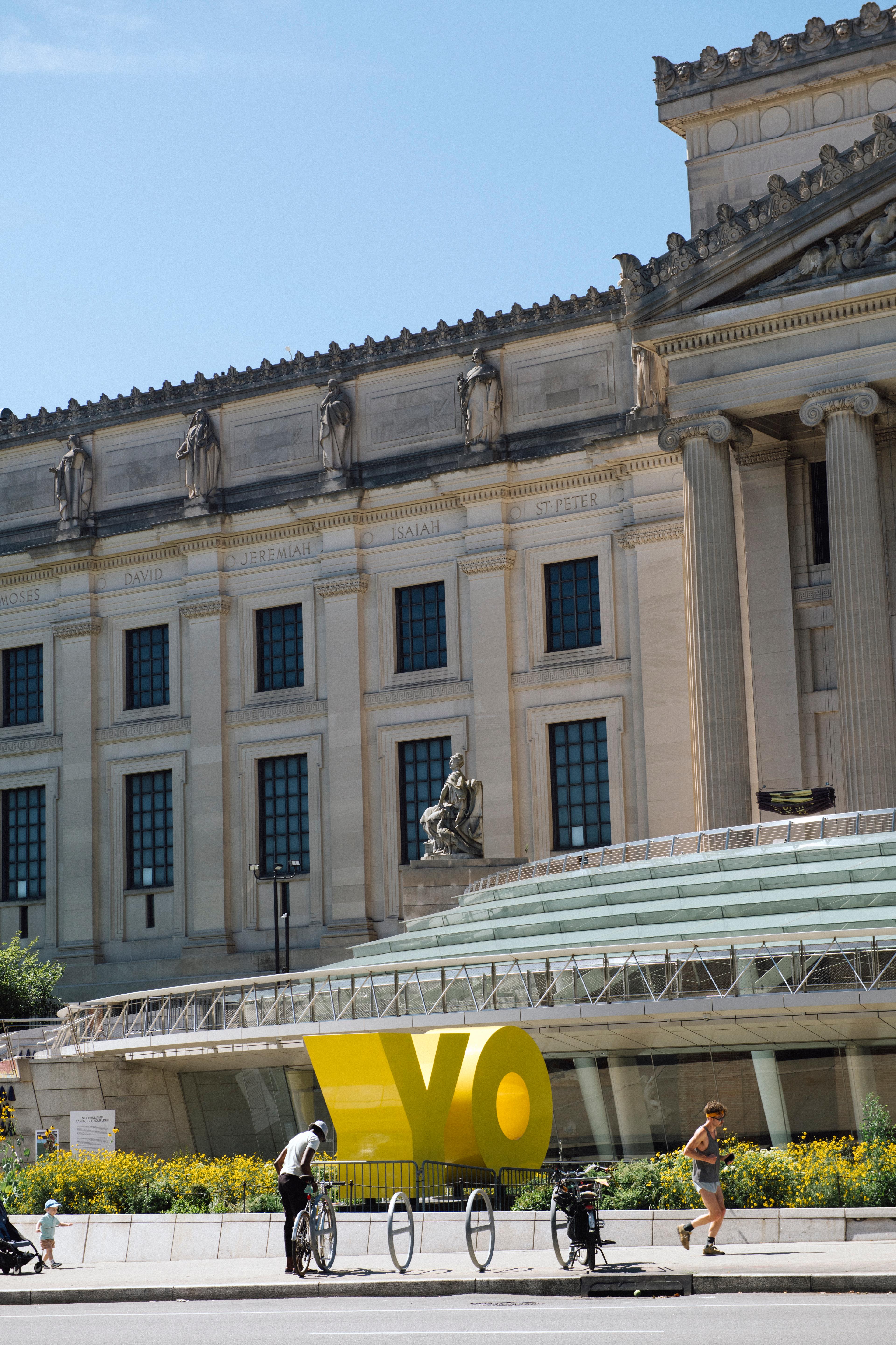 Detail of a Museum plaza featuring visitor locking up a bike in front of a large sculpture that reads "YO"